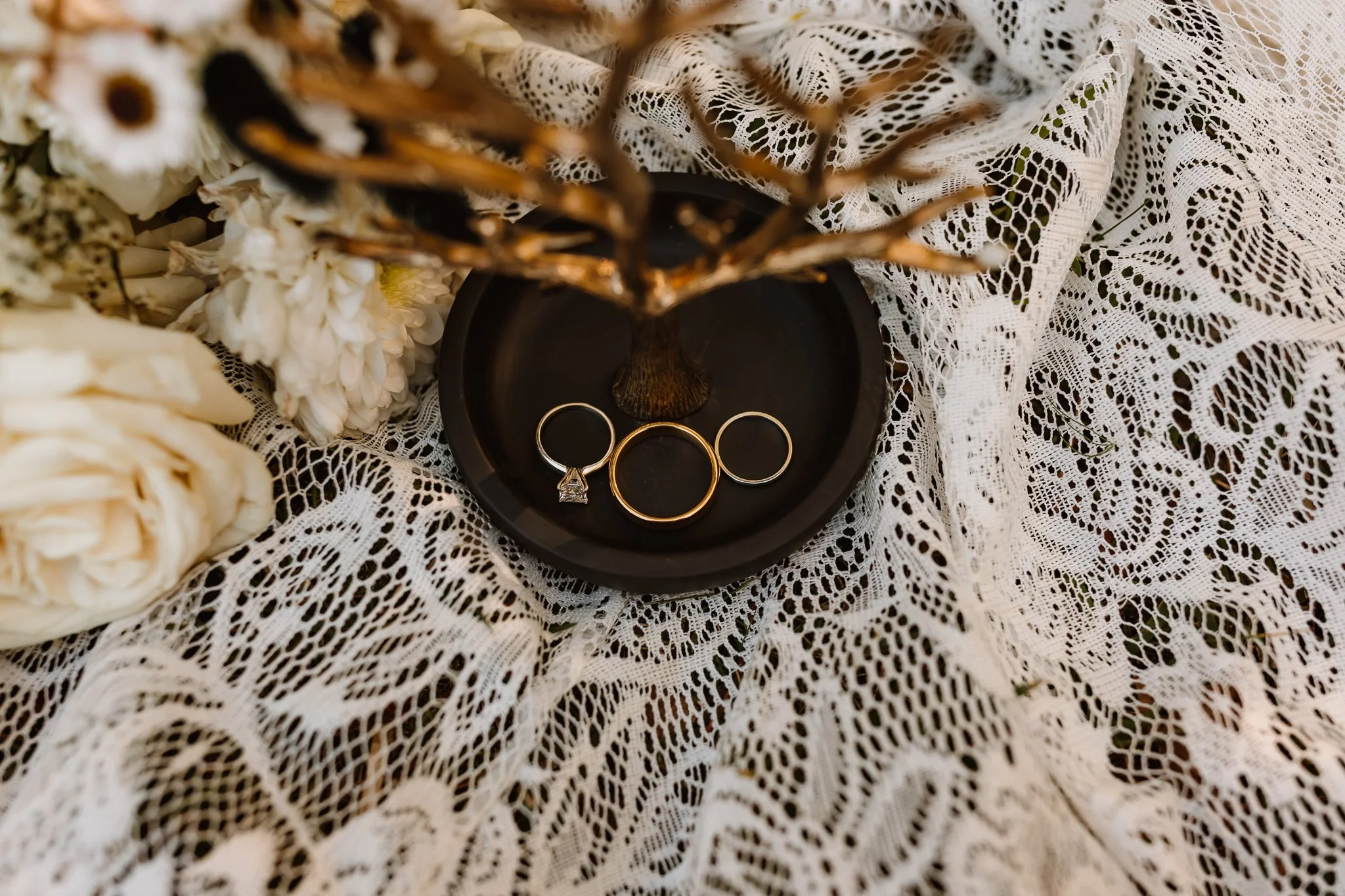 Three rings, including a wedding band and an engagement ring, placed on a black dish. The dish is on white lace fabric, with a decorative tree and white flowers around it.