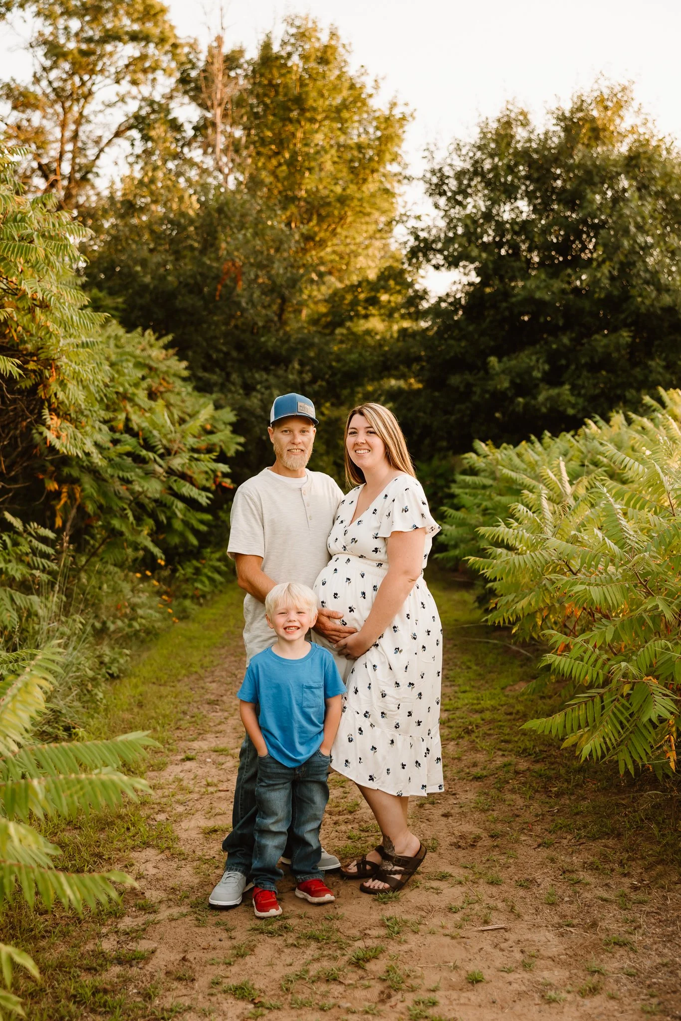 A family of three standing on a dirt path surrounded by green trees and plants during sunset. The woman is pregnant, wearing a white dress, and stands in the middle. The man is on her left, wearing a baseball cap and a light-colored t-shirt. A young 