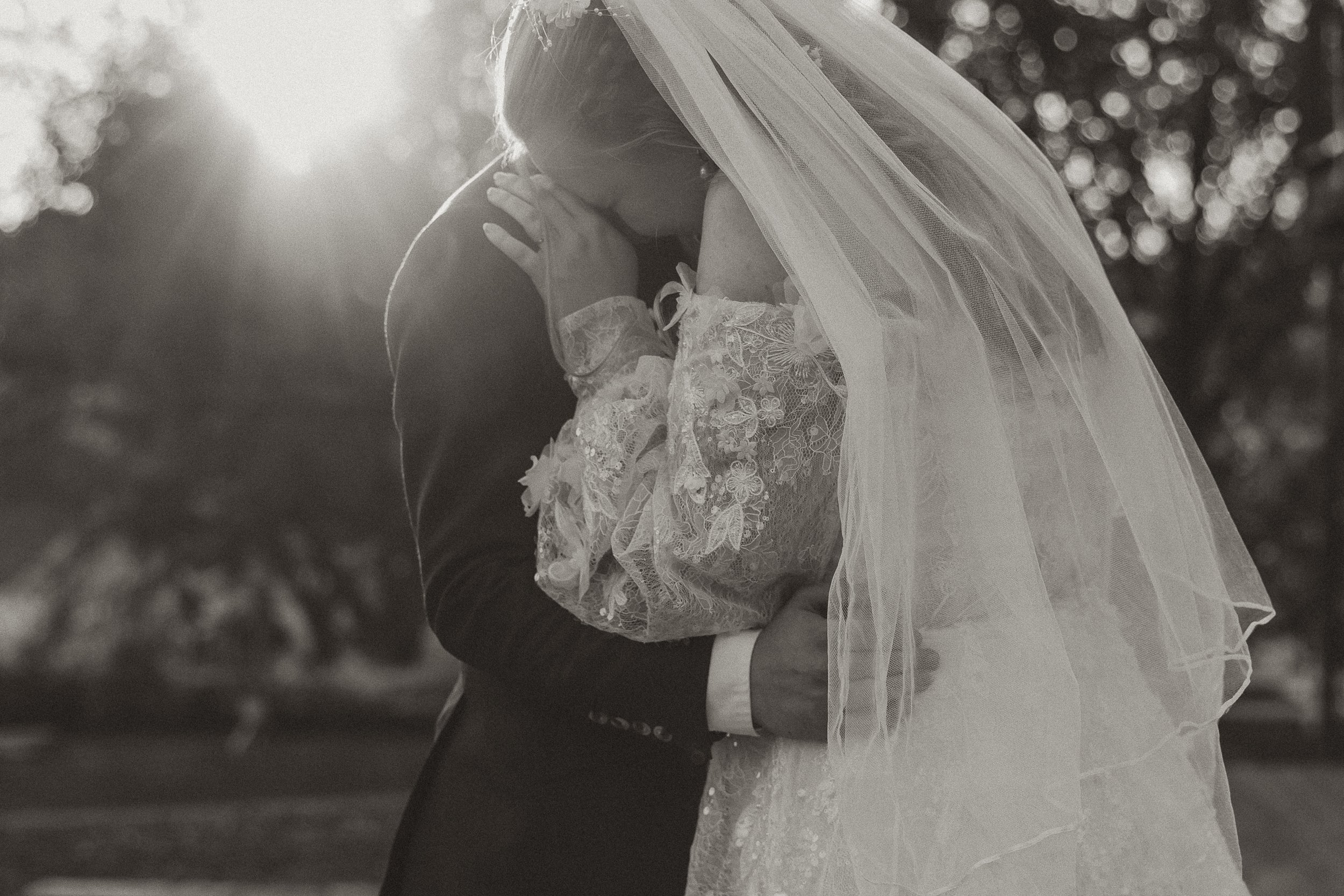 A black-and-white photo of a bride and groom kissing outdoors during sunset, with the bride wearing a lace wedding dress and veil, and the groom in a suit.