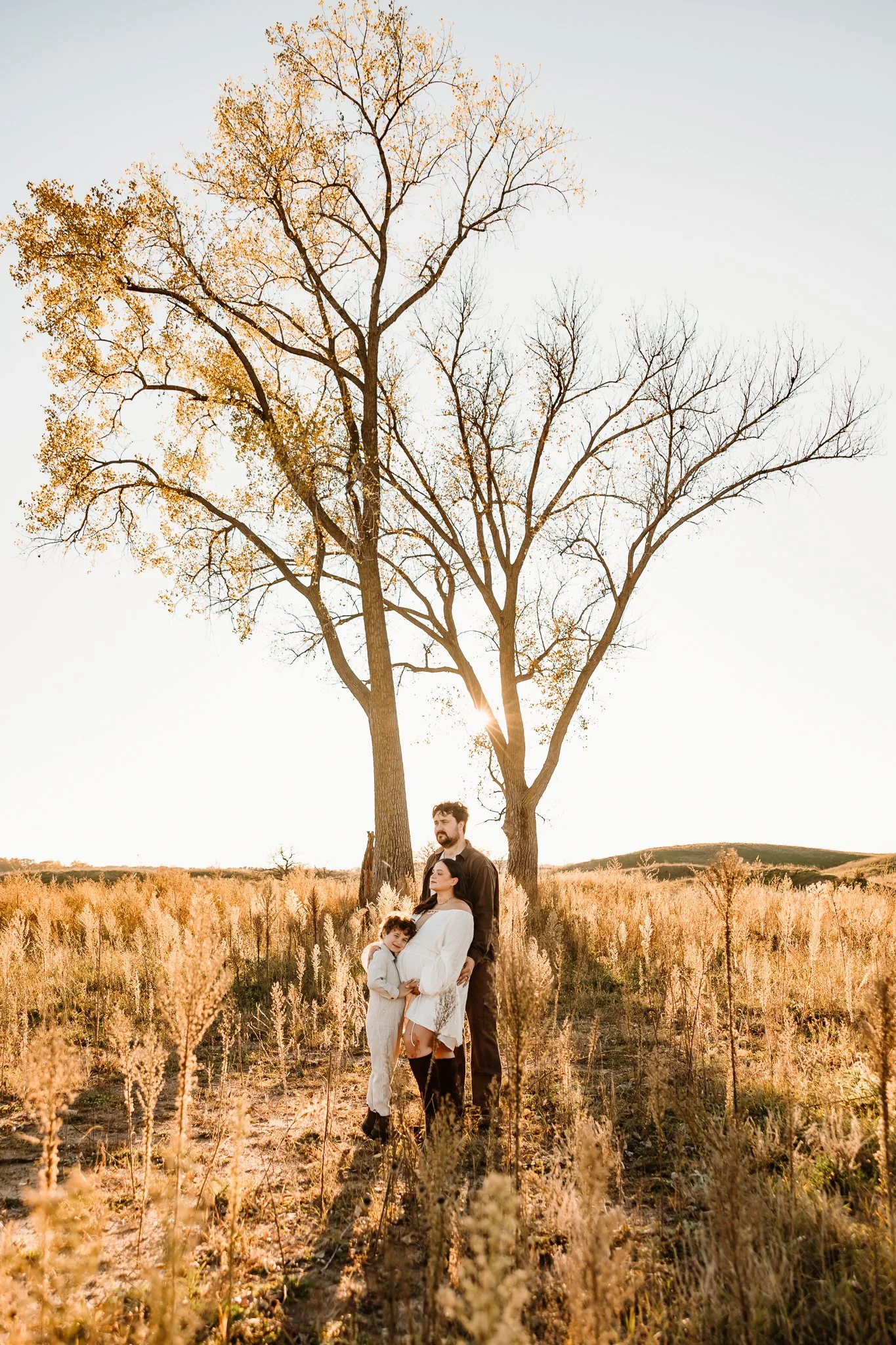 A family of three standing outdoors in a field with tall, dry grass during sunset, with two large leafless trees in the background and the sun shining through the branches.