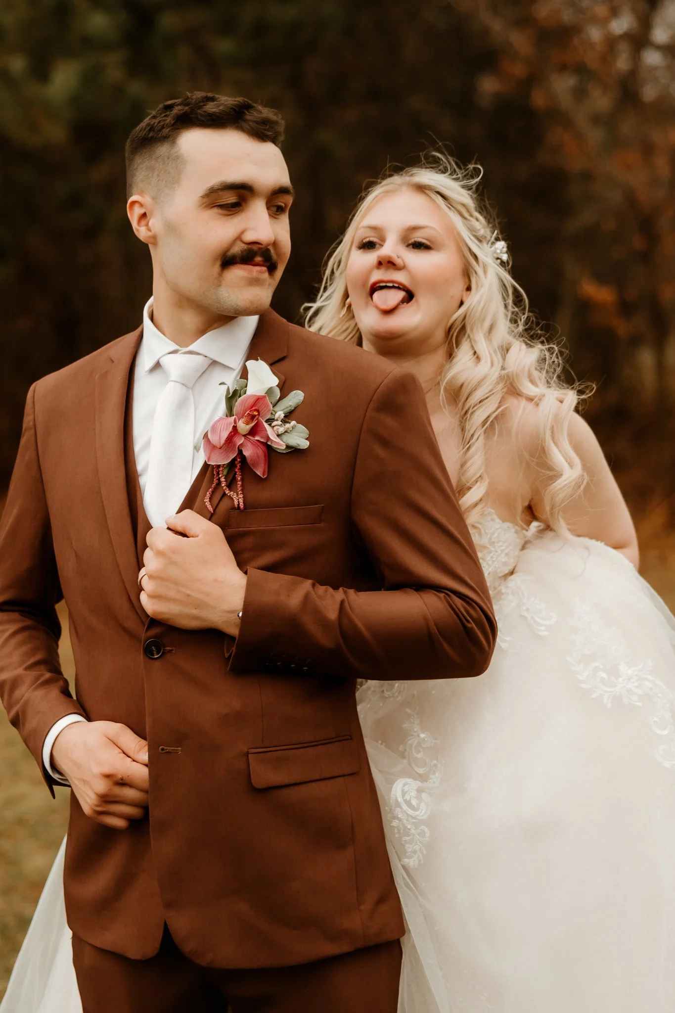A bride and groom outdoors during autumn. The groom is in a brown suit with a floral boutonniere, and the bride is in a white wedding dress, making a playful face with her tongue out behind the groom.