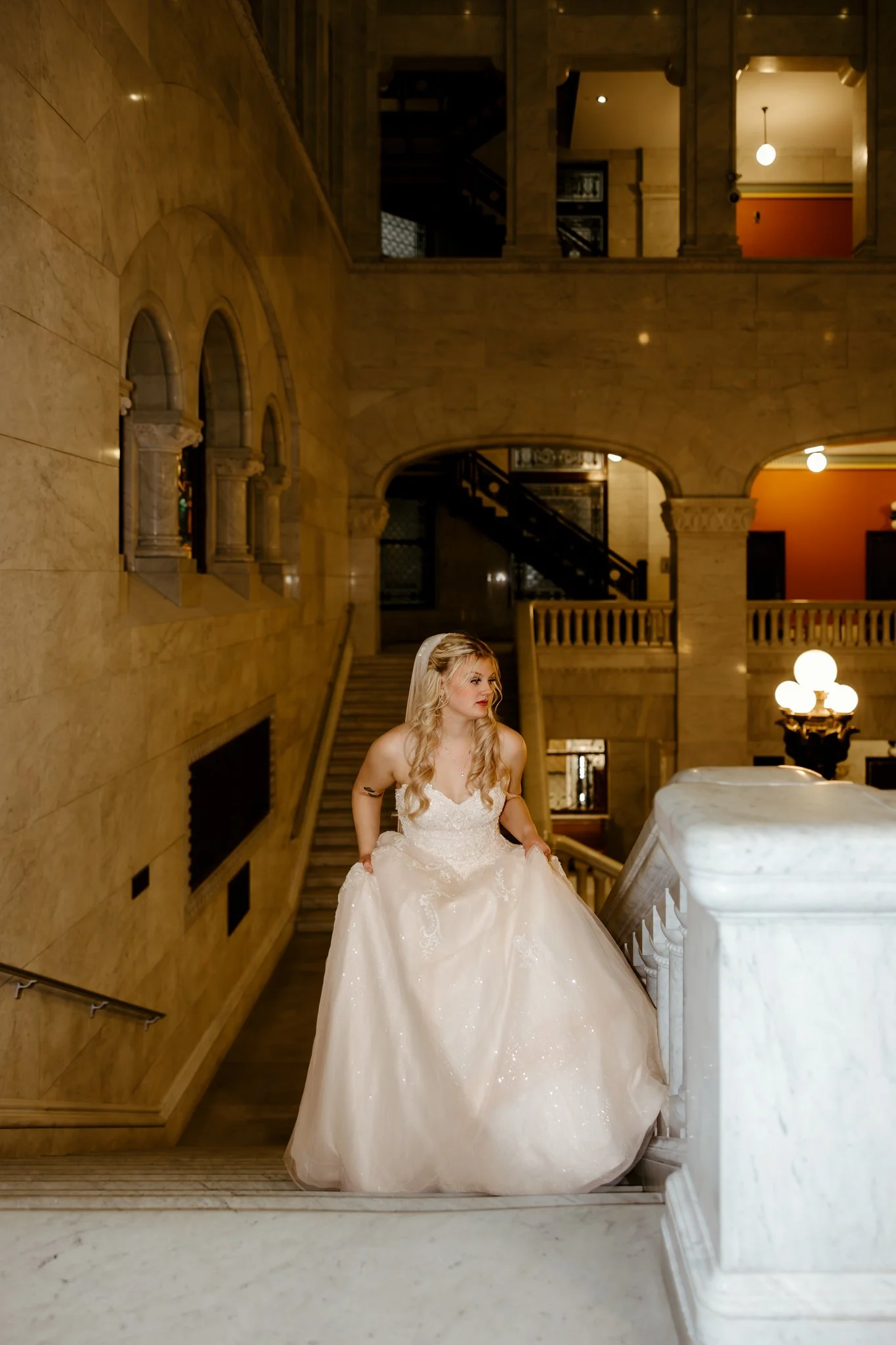 A bride in a white wedding gown standing on a staircase inside a historic building with marble walls and arches.