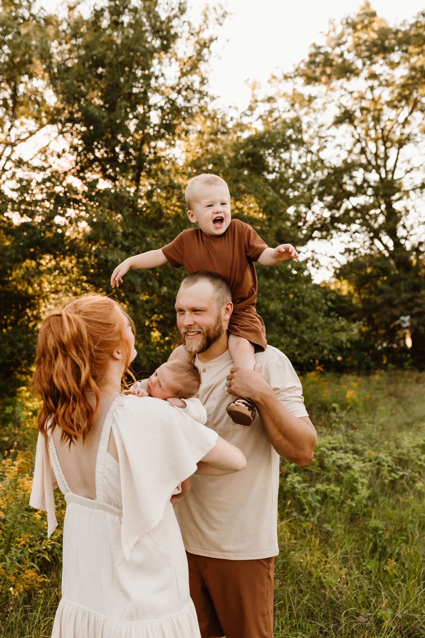A family of four enjoying outdoors during golden hour. The father, with a beard, is carrying a young girl on his shoulder, who is smiling and wearing a brown shirt. The mother, with red hair, is holding a newborn baby close to her chest. The backgrou