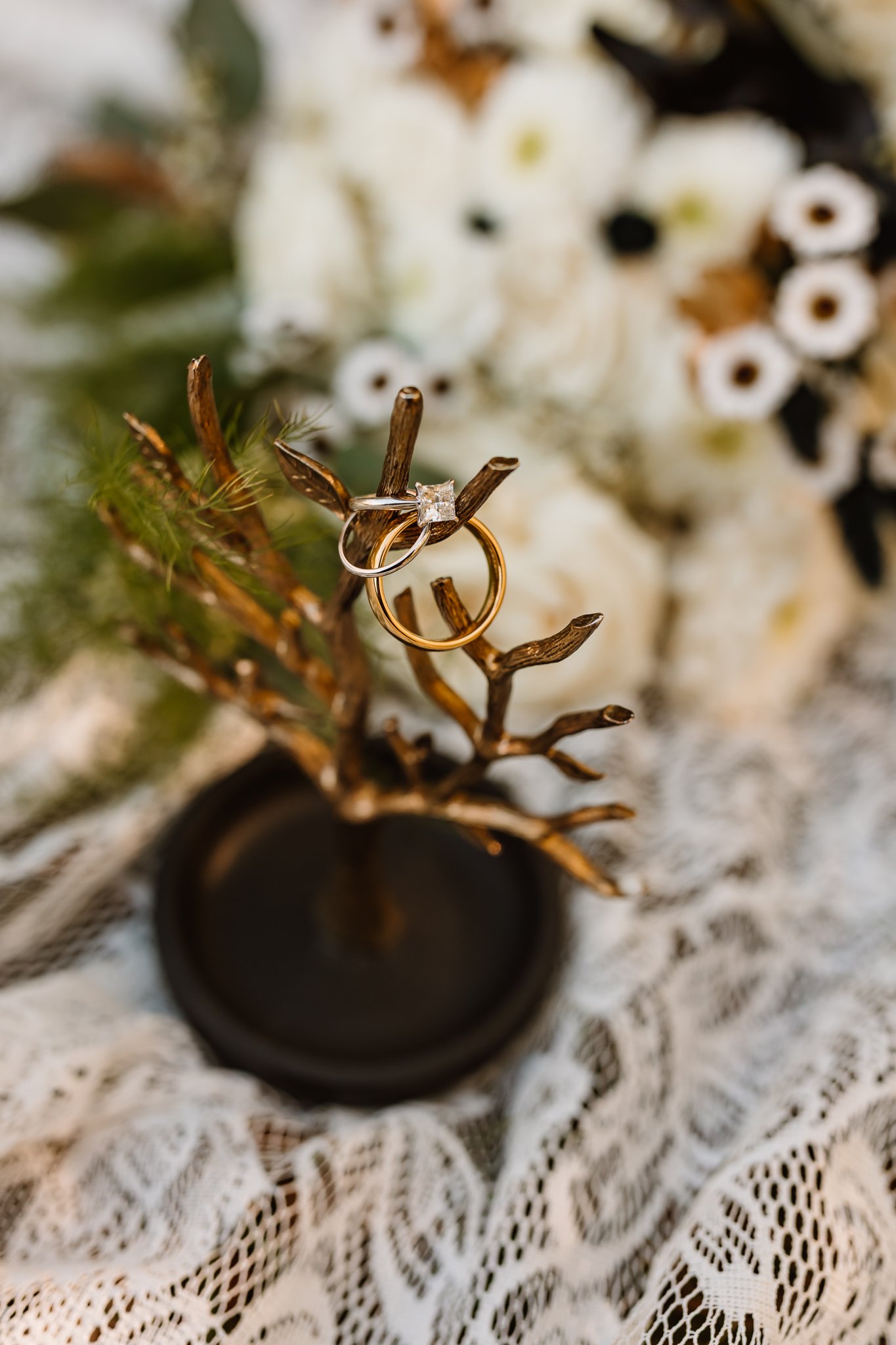 Wedding rings, one with a diamond, resting on a small decorative gold branch, with a floral bouquet in the background on lace fabric.