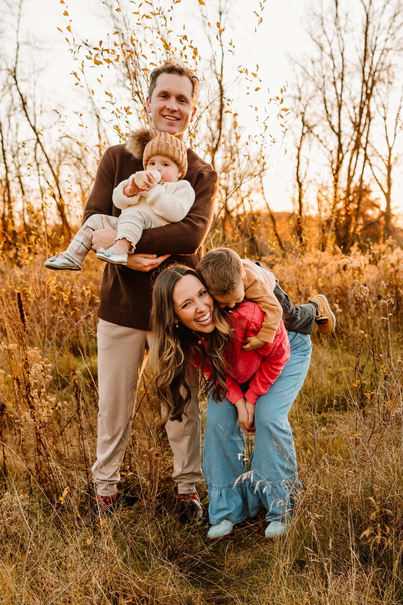 A family of four enjoying fall outdoors in a field with trees and golden leaves, two adults and two children, one of whom is being held and the other is being playfully lifted.