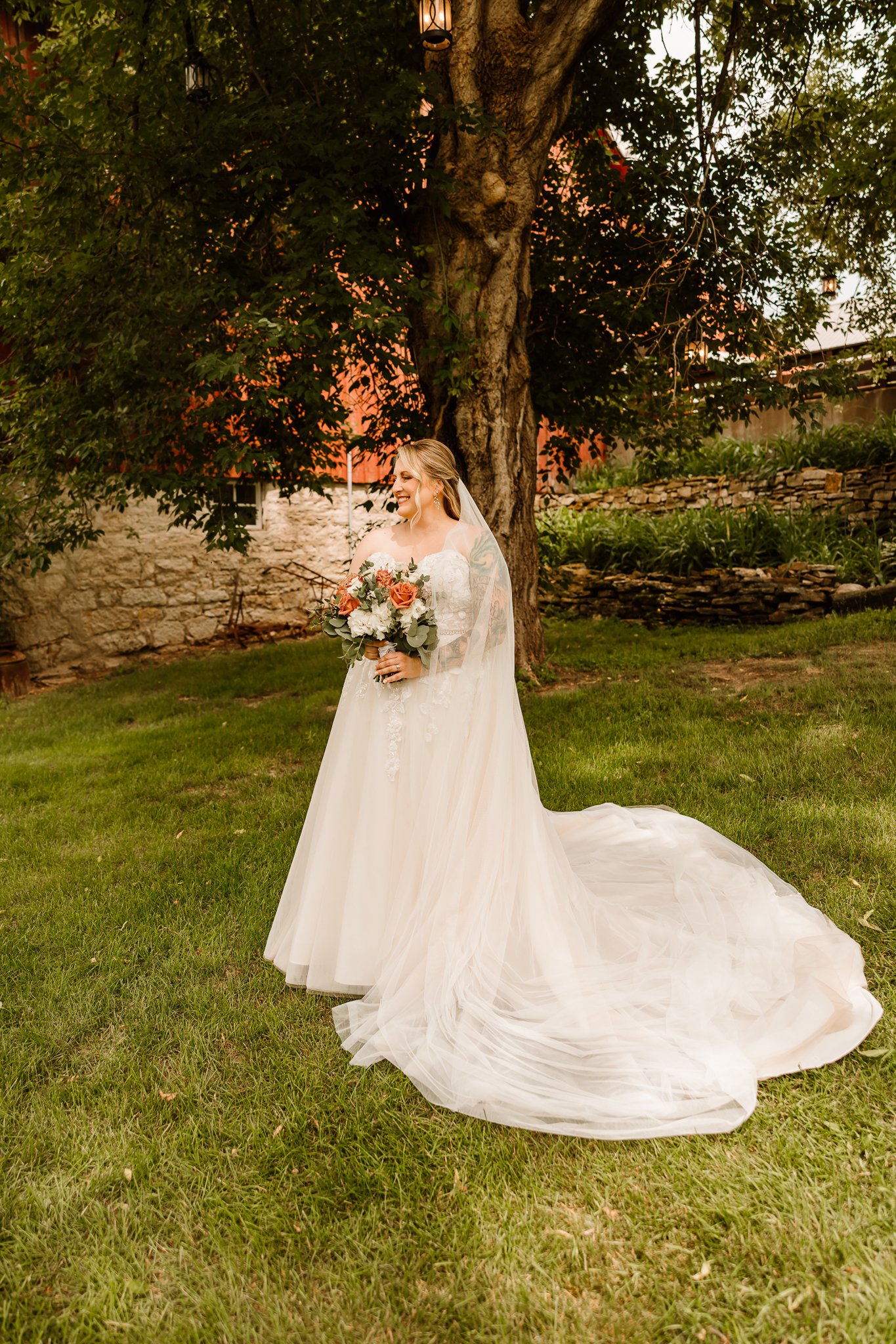 A bride in a white wedding gown holding a bouquet of pink and white flowers, standing on grass near a large tree with green foliage.