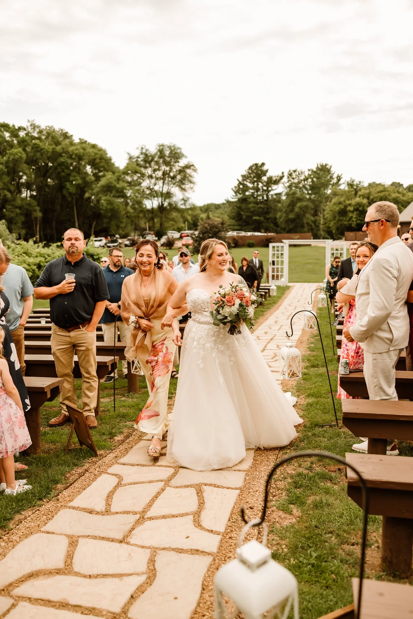 Bride walking down the aisle with a woman, possibly her mother, smiling beside her. Guests standing and sitting on benches on either side, outdoors at a wedding ceremony.
