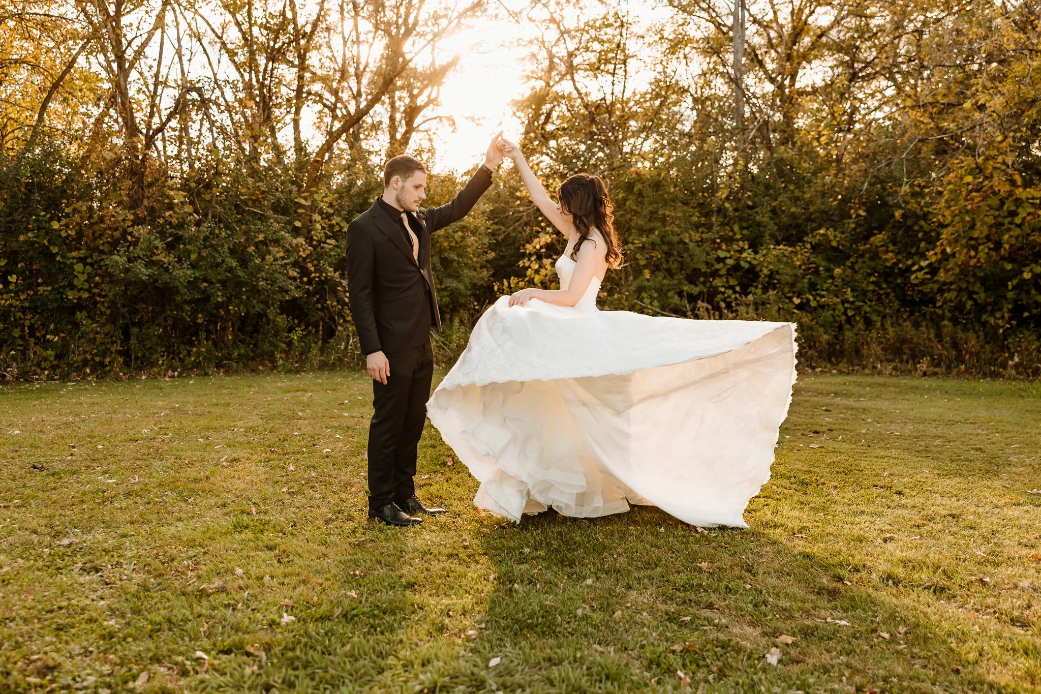 A wedding couple dancing outdoors on grass during sunset, with trees in the background.