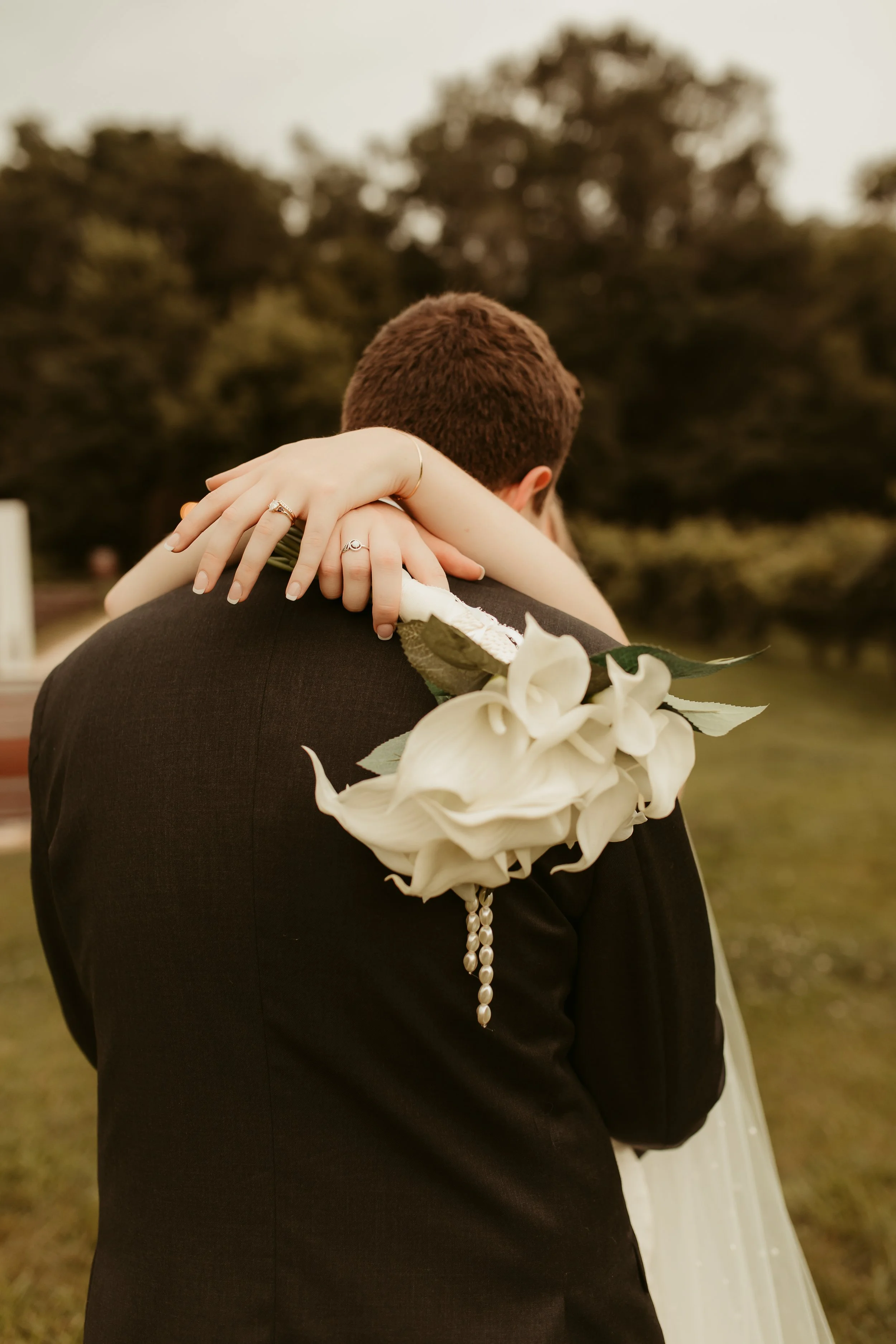 A couple sharing an embrace outdoors, with the woman's hands on the man's back, holding a bouquet of white flowers. The woman is wearing rings and a bracelet, and the man is in a black suit. Trees are visible in the blurred background.