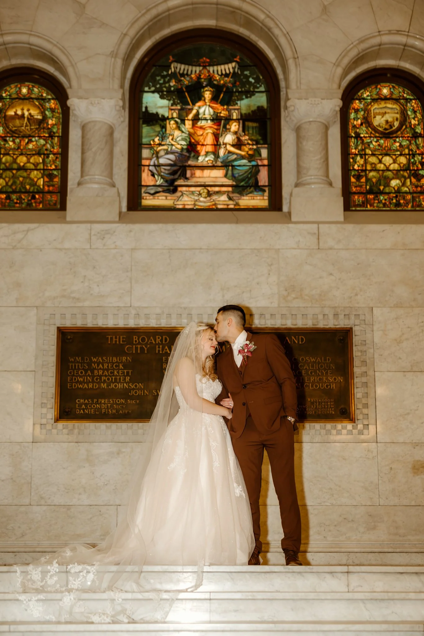 A bride and groom kissing in a courthouse with stained glass windows behind them.