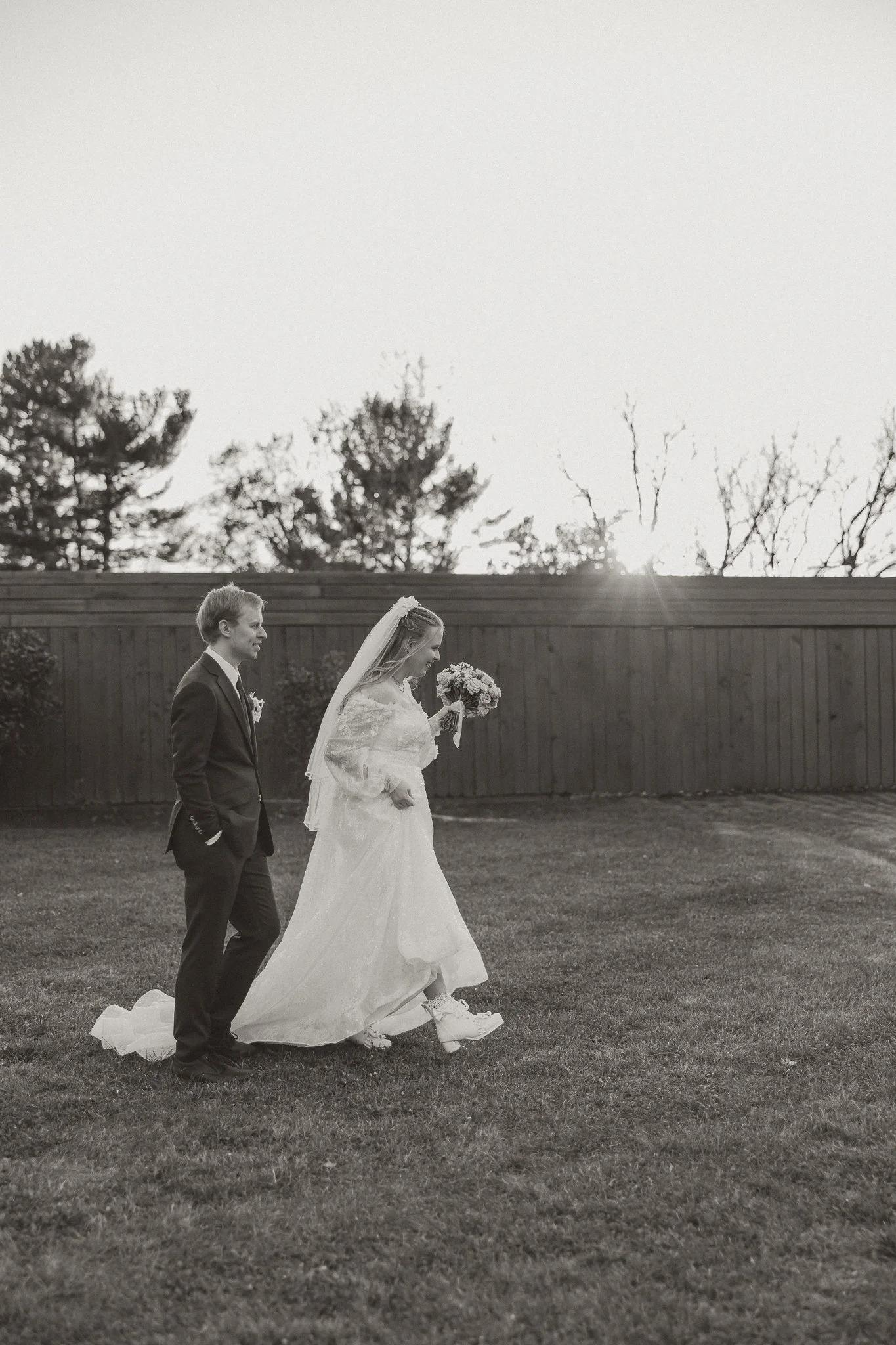 Black and white photo of a bride and groom walking on grass outdoors, with trees and a fence in the background. The bride is holding a bouquet and wearing a long dress and veil. The groom is in a suit with his hands in his pockets.