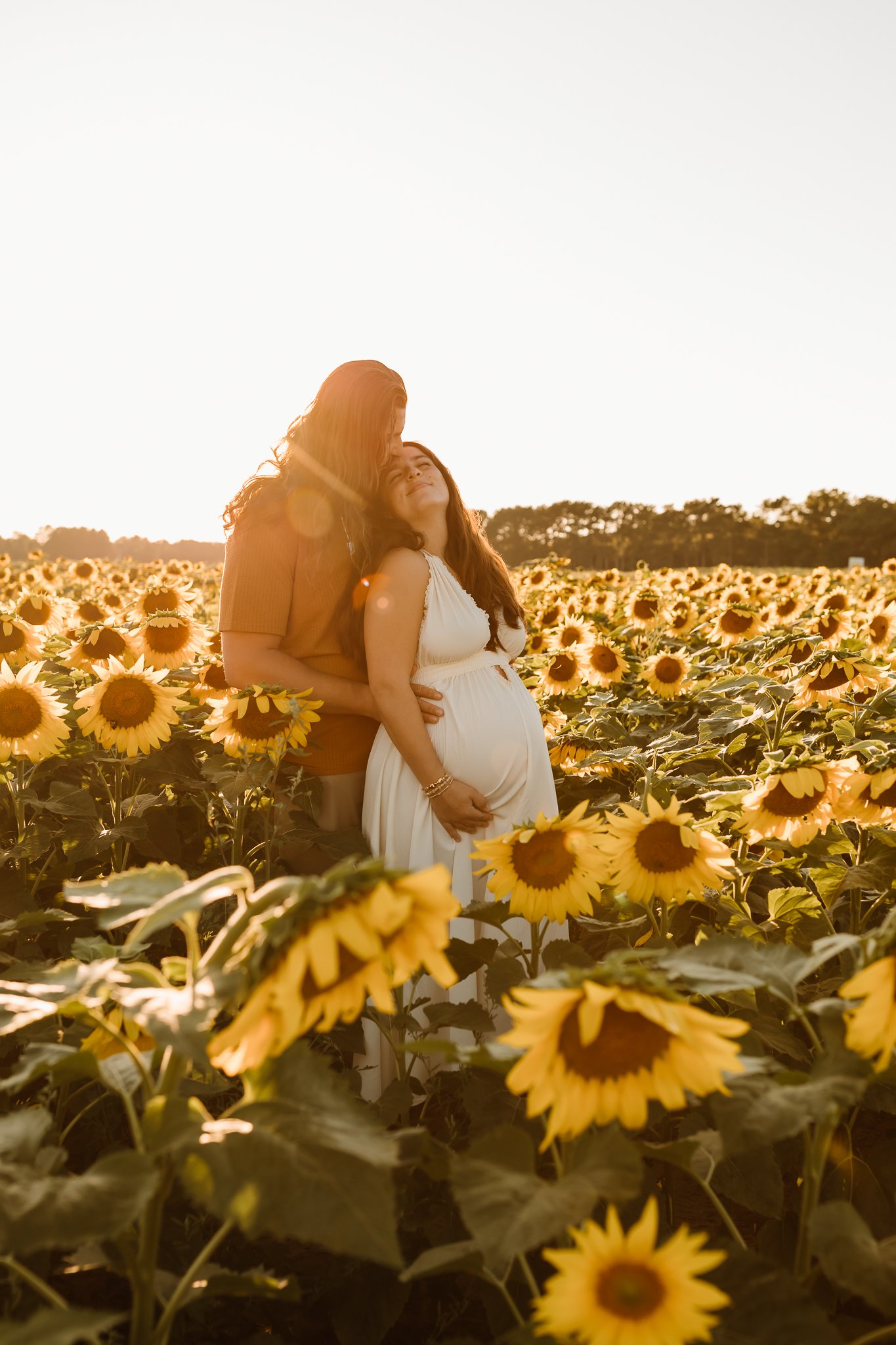 A couple standing in a field of sunflowers during sunset, with the woman appearing pregnant and smiling, and the man embracing her.