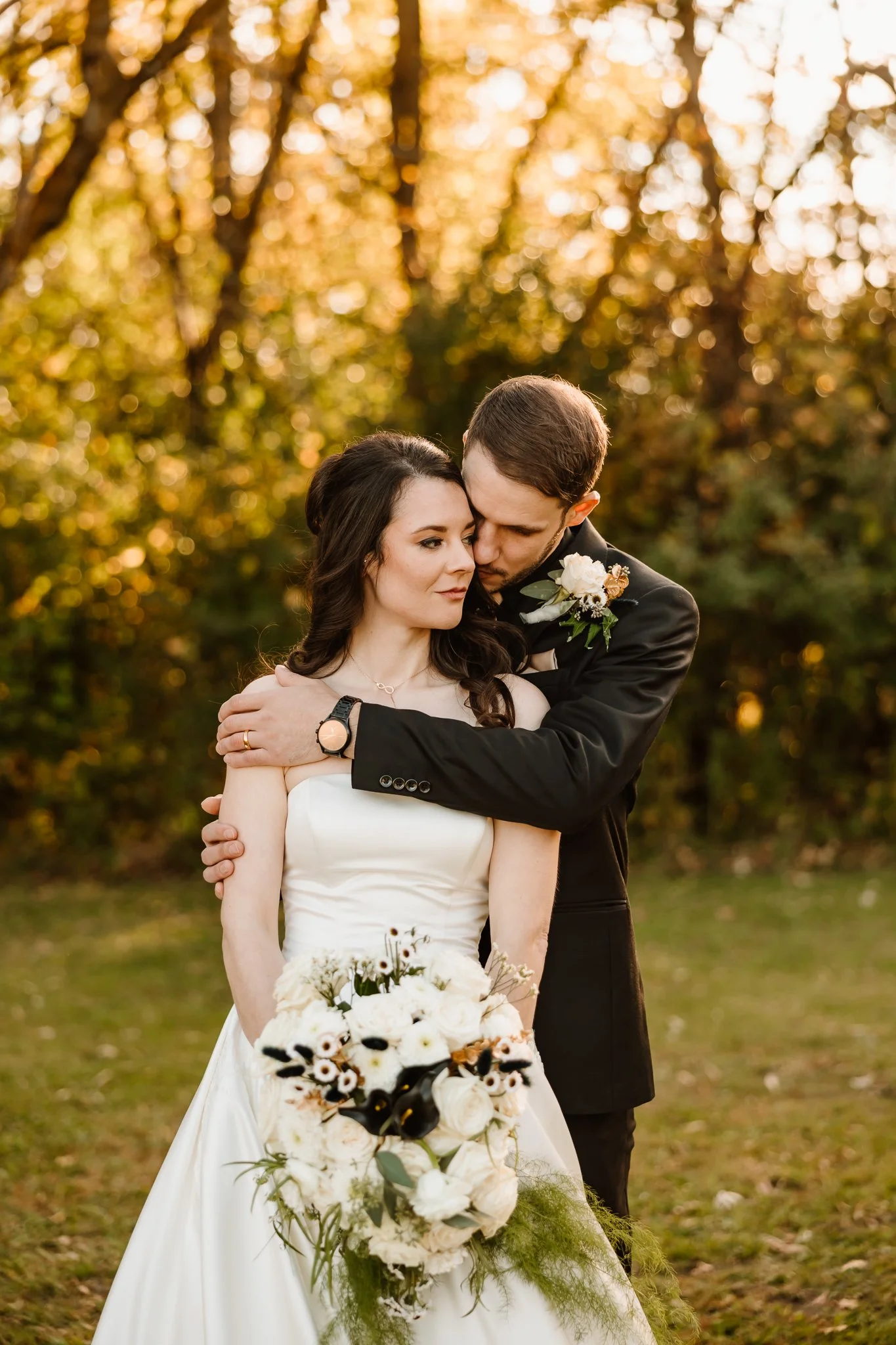 A bride and groom embrace outdoors during sunset, the bride holding a bouquet of white and dark flowers, surrounded by autumn trees.