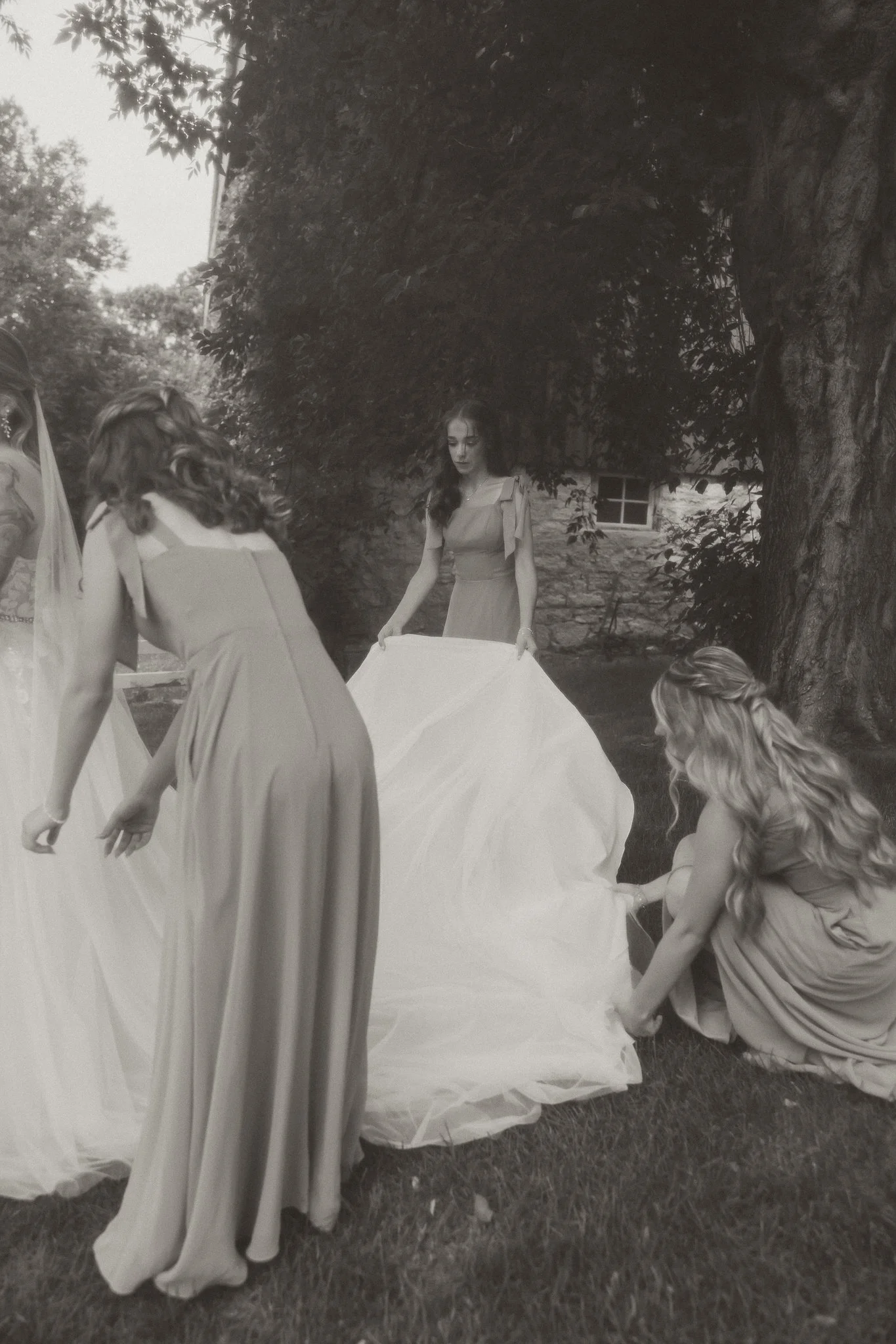 Three women preparing a wedding dress outdoors near a large tree and a rustic building.