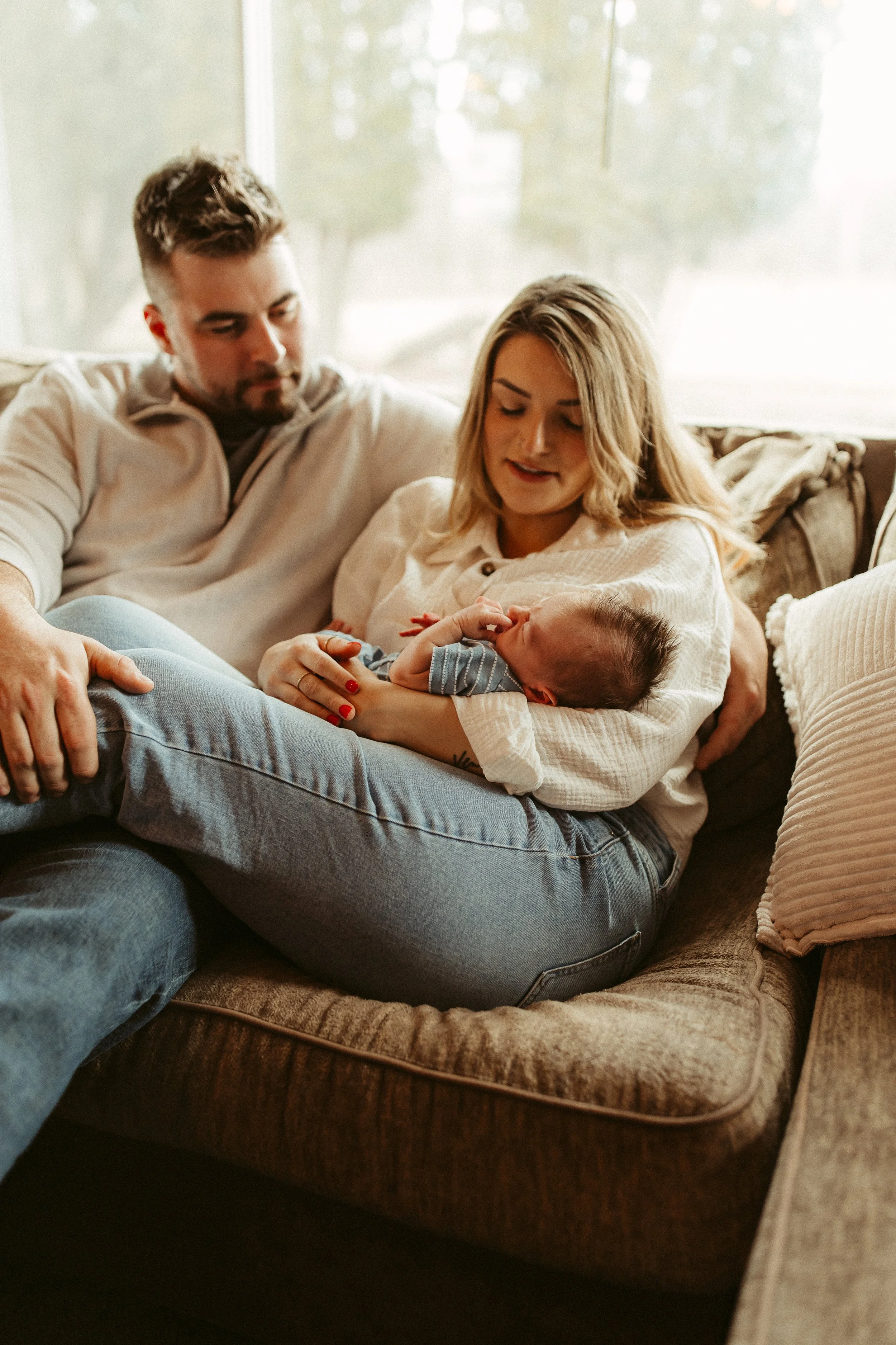 A young woman is sitting on a couch, cradling a sleeping baby in her lap while a man sits beside her, all looking at the baby.