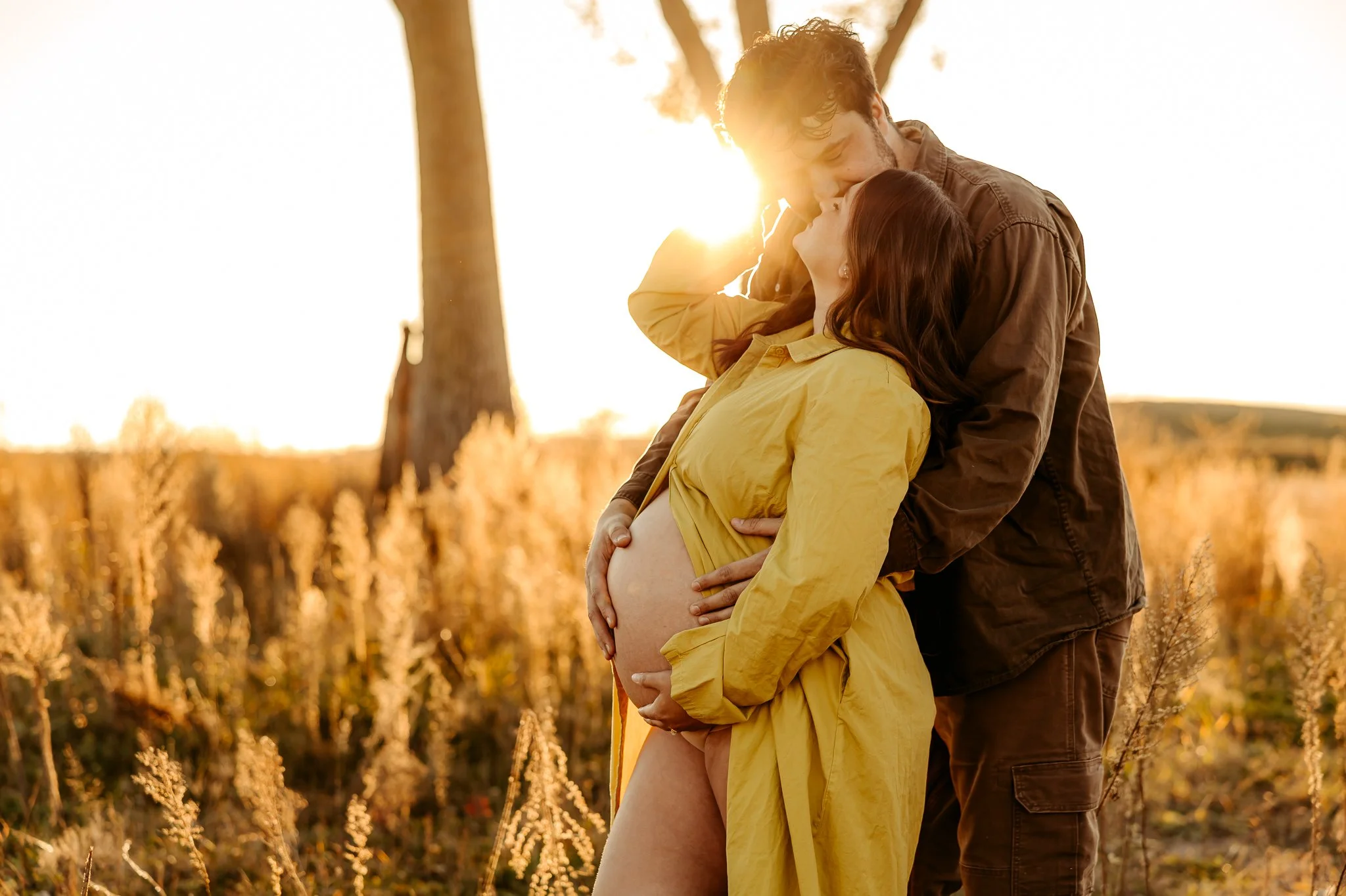 A pregnant woman in a yellow dress and a man are sharing a romantic kiss outdoors during sunset, surrounded by tall grass and a tree.