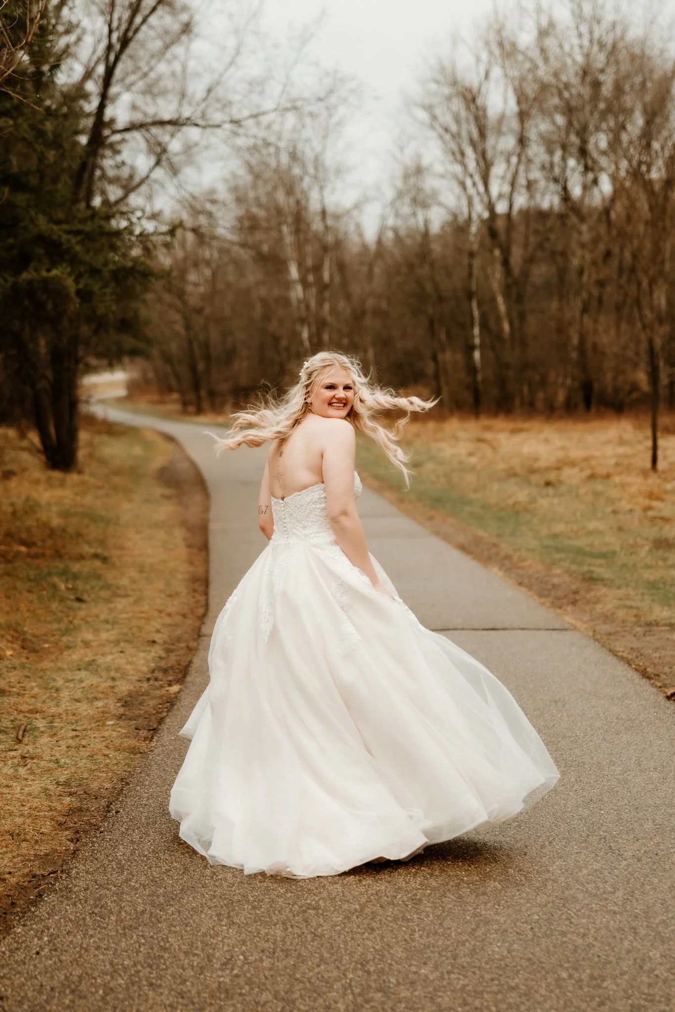 A blonde woman in a white wedding dress spinning on a paved path in a leafless park during autumn or winter.
