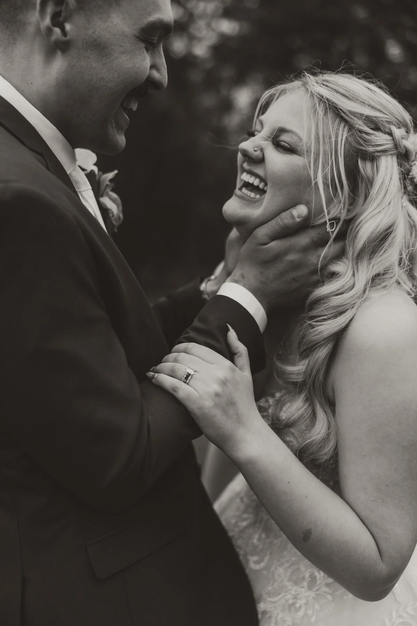 Black and white photo of a happy couple, likely at their wedding, with the man gently holding the woman's face as they smile and laugh together outdoors.