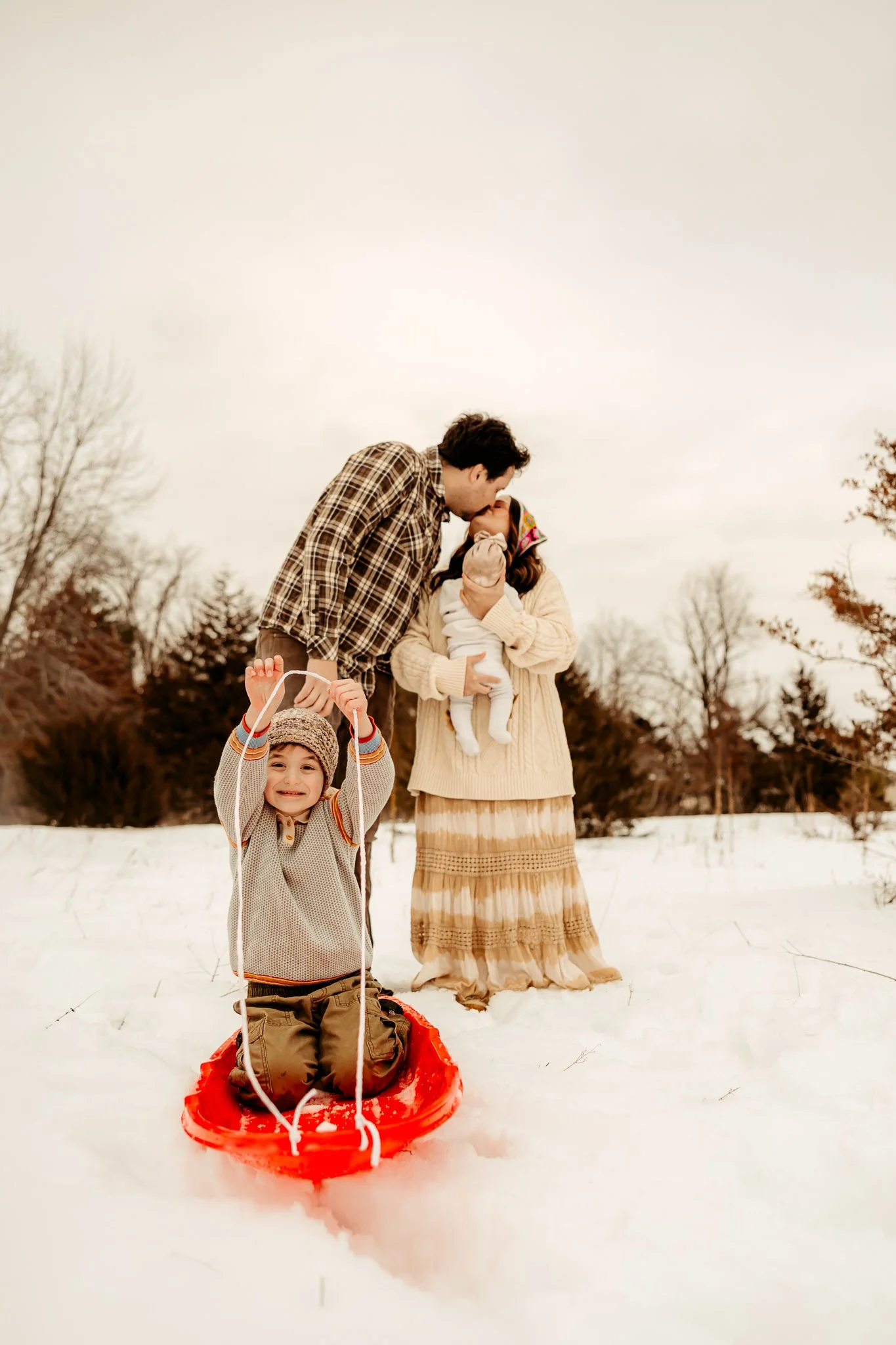 A family of four outdoors in the snow, with two parents, a young boy on a sled, and a woman holding a baby, sharing a kiss.