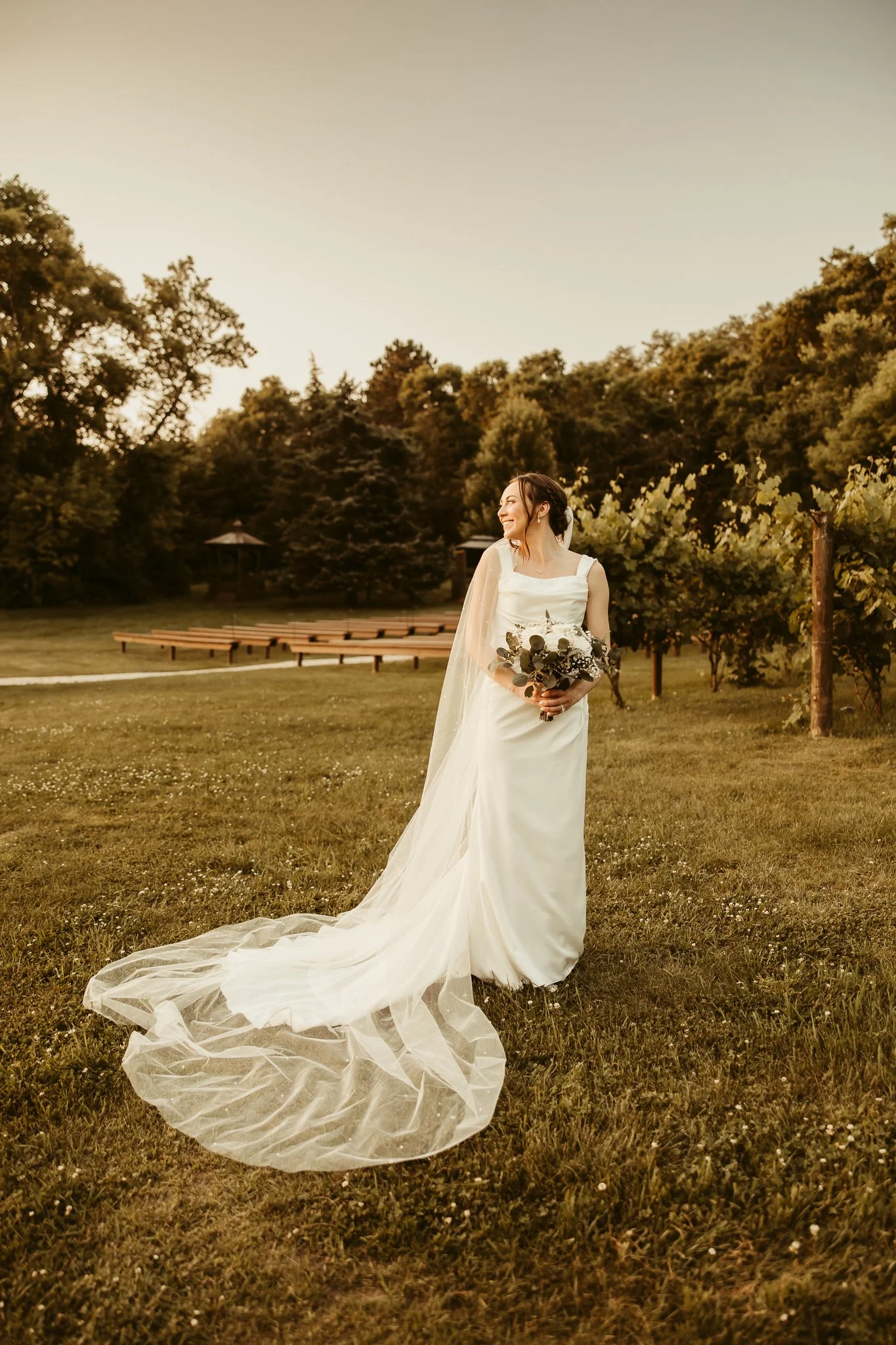 Bride in a white wedding dress holding a bouquet, standing on grass in a scenic outdoor setting during sunset.