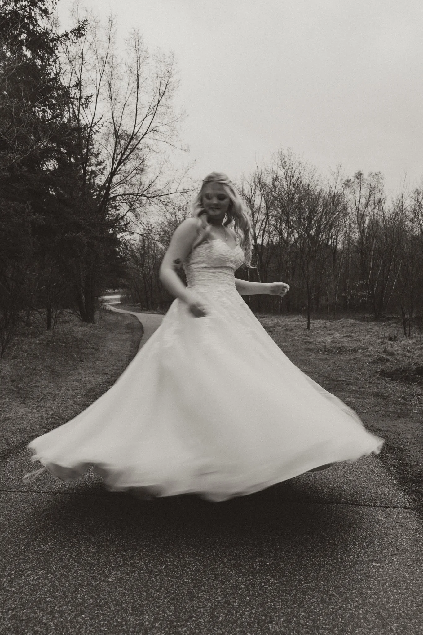 A woman in a wedding dress spinning on a road in a rural area with bare trees, captured in black and white.