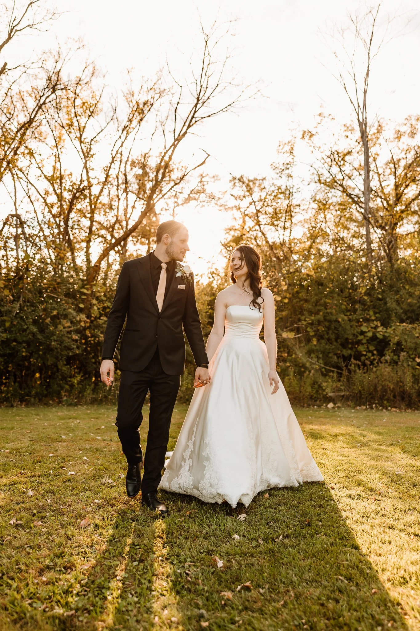 A bride and groom walk hand in hand outdoors during sunset, surrounded by trees and grass.