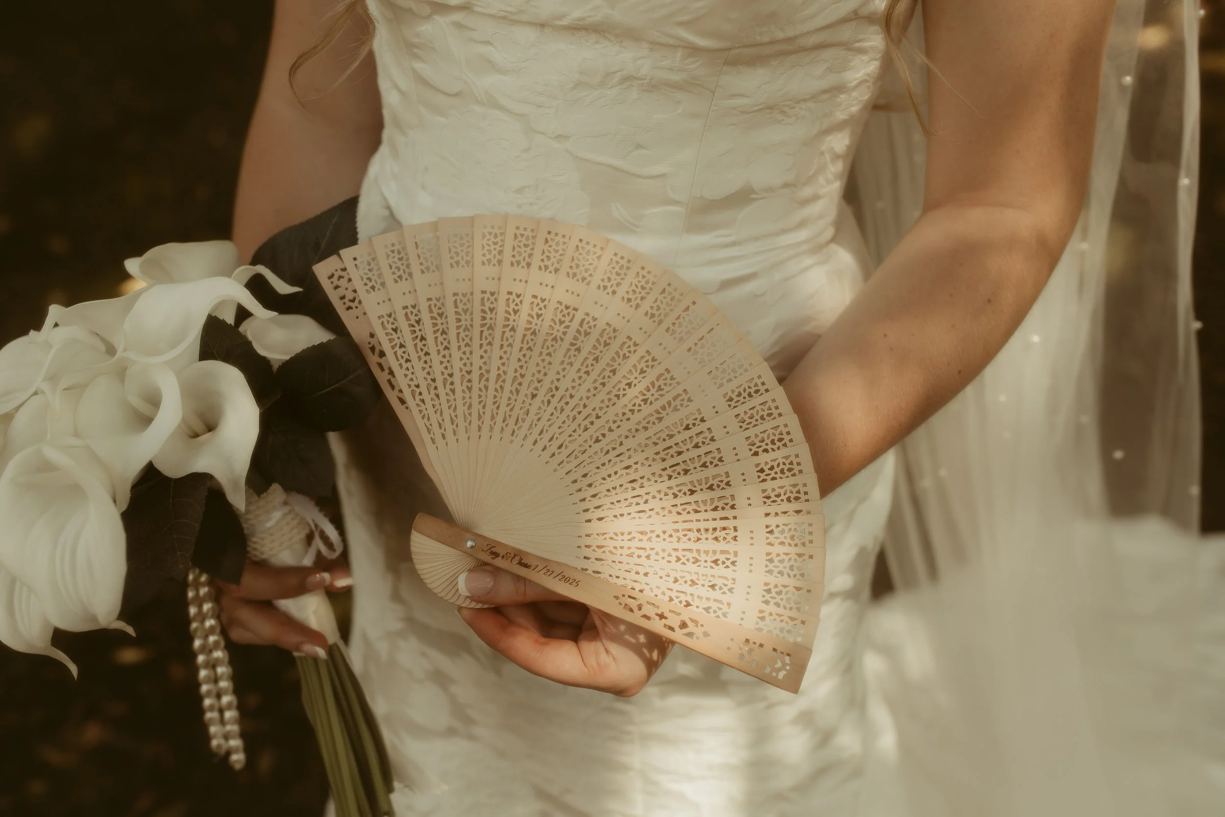 A woman in a white wedding dress holding a beige decorative hand fan and a bouquet of white calla lilies with dark leaves and pearl accents.