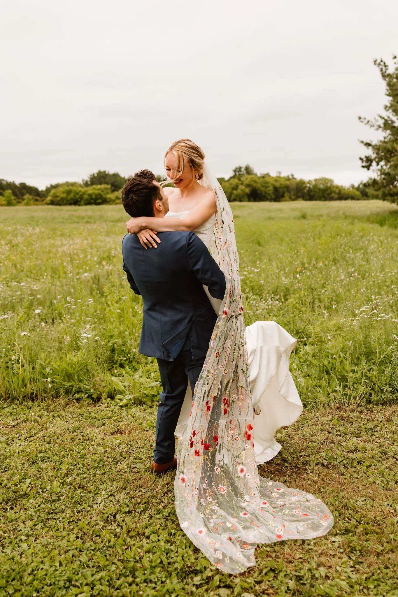 A bride and groom embrace outdoors in a grassy field during their wedding, with the bride sitting on the groom's lap and both smiling.