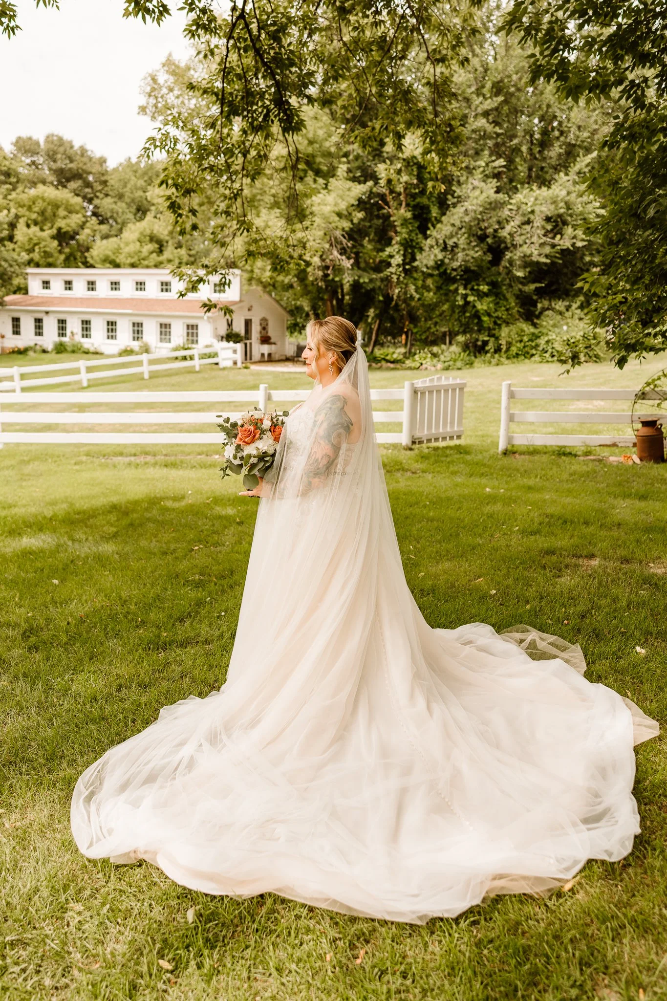 A bride in a white wedding gown with a long veil, holding a bouquet of flowers, standing outdoors on a grassy area with trees and a white fence in the background.