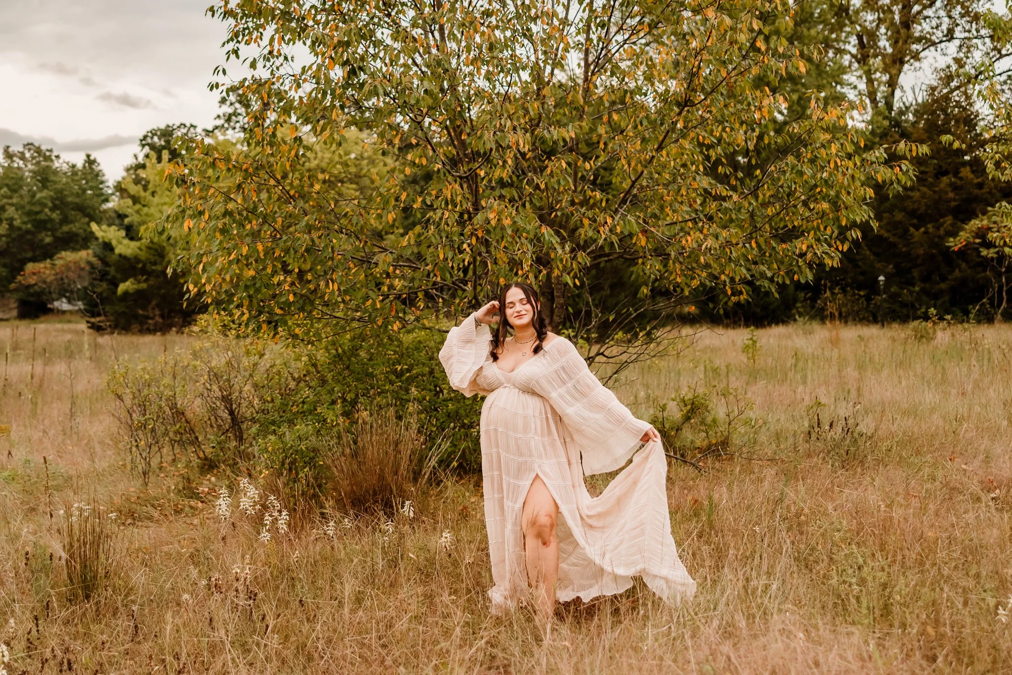 A woman in a flowing, light-colored dress standing in a field with tall grass and a large tree in the background. She is posing with a smile, holding part of her dress, with autumn-colored trees behind her.