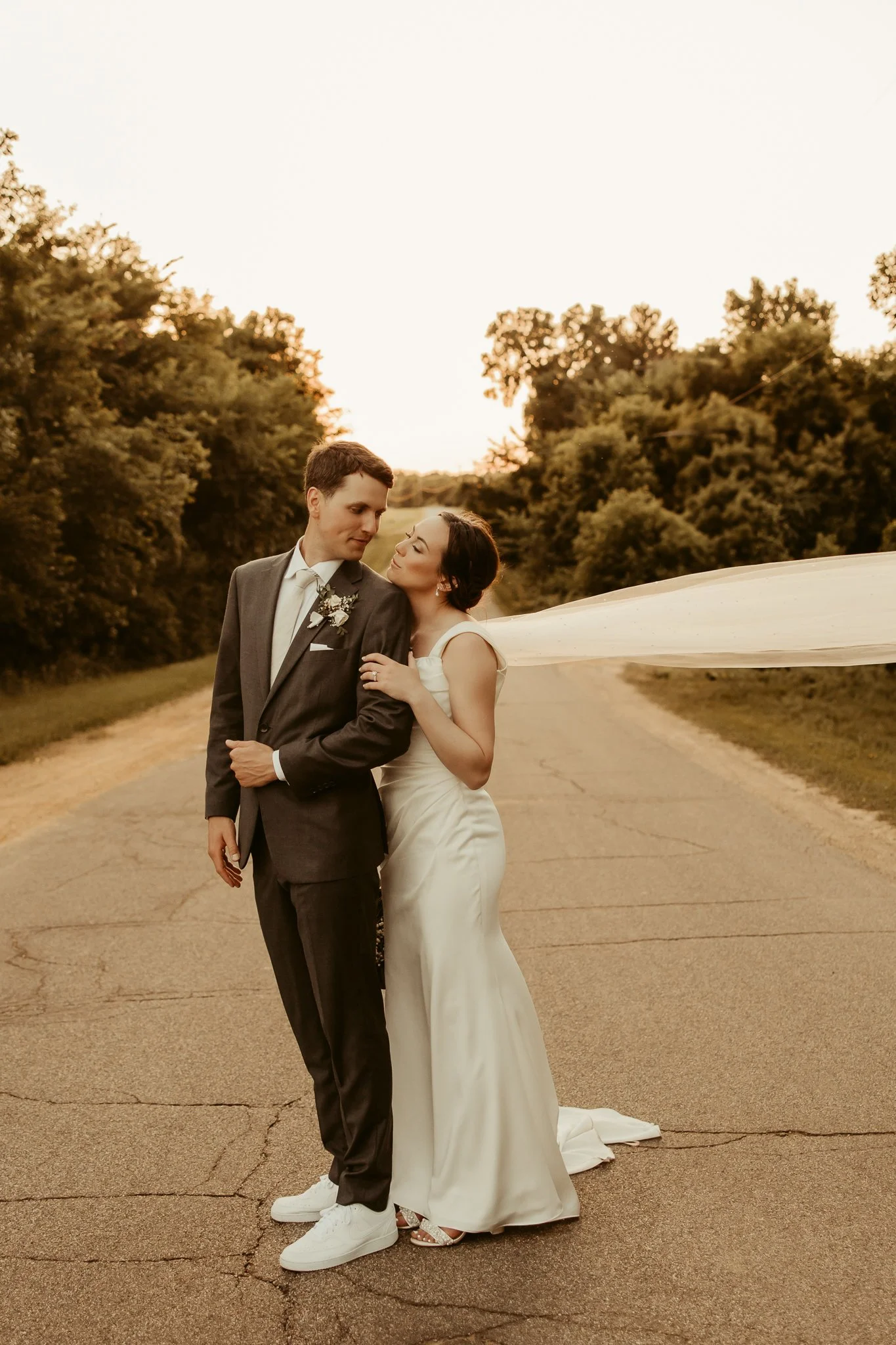 A newlywed couple in wedding attire standing on an empty road during sunset, surrounded by trees.