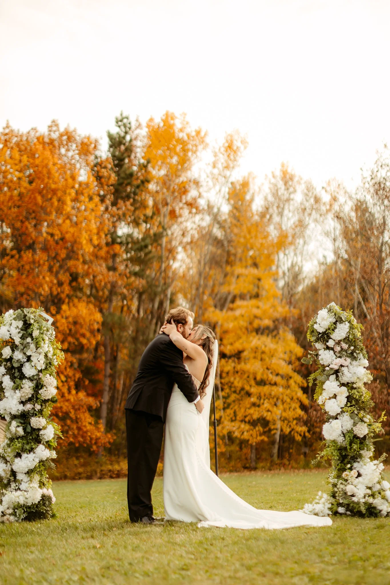 A bride and groom sharing a kiss during their outdoor wedding ceremony, surrounded by fall foliage and floral arrangements.