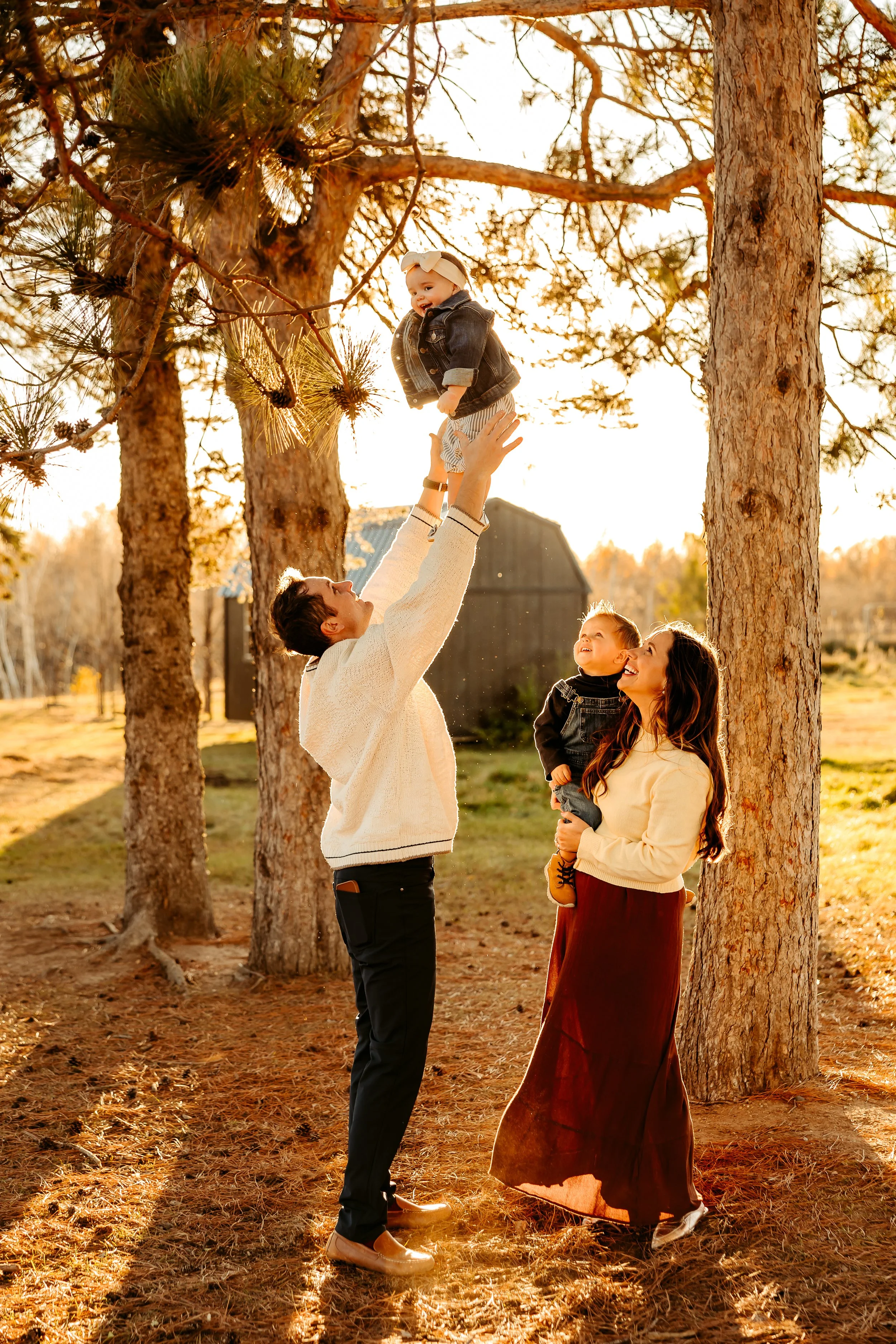 Family outdoors in a forest during sunset, playing with a young child in a tree.