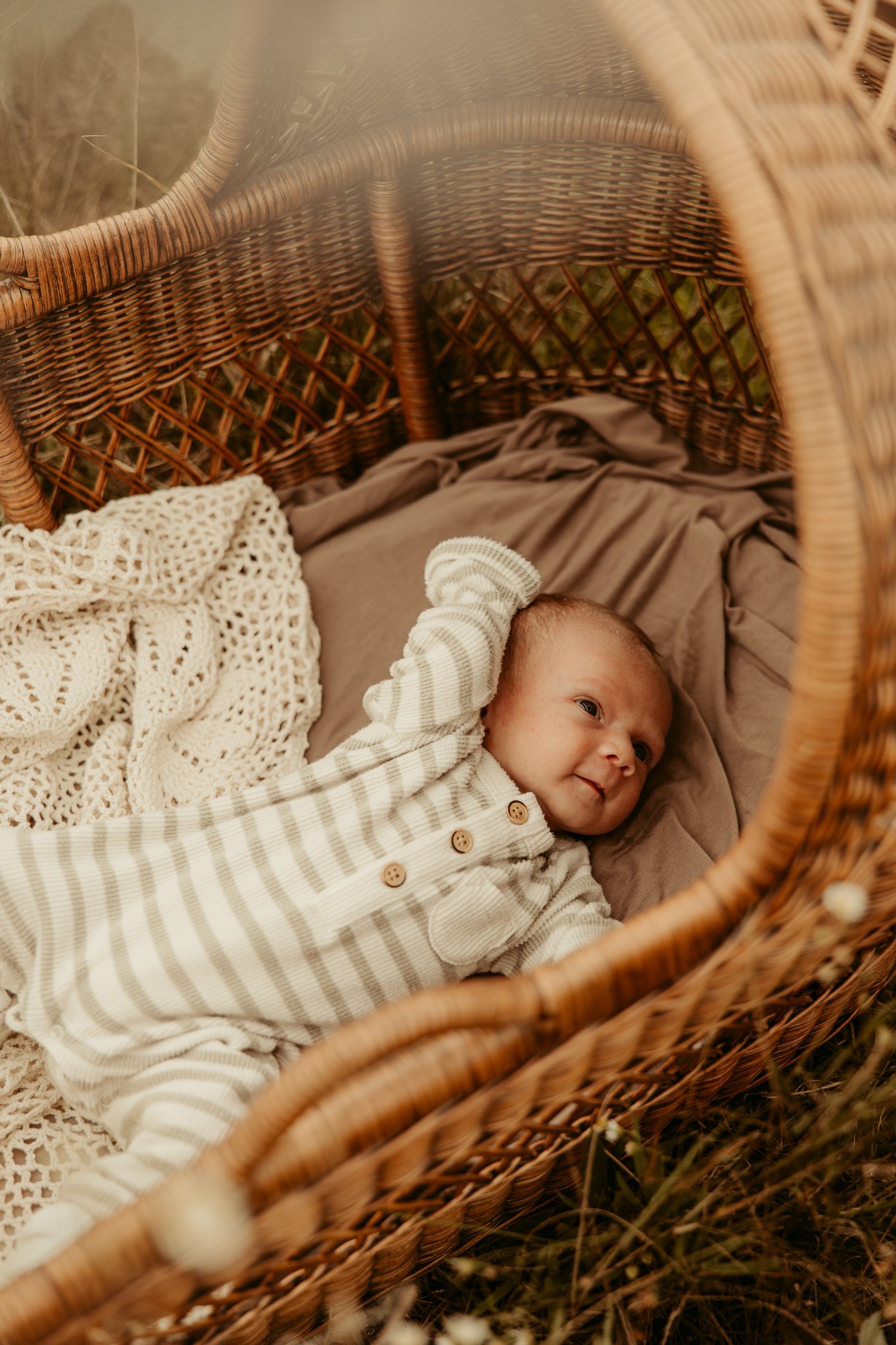 A baby lying in a wicker bassinet outdoors, smiling and looking to the side, wearing a striped beige and white onesie.