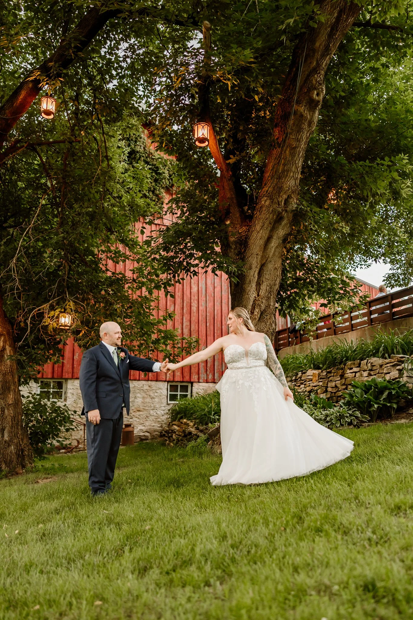 Bride and groom holding hands outdoors during wedding ceremony, standing on grass near trees and a red barn, with hanging lantern lights.