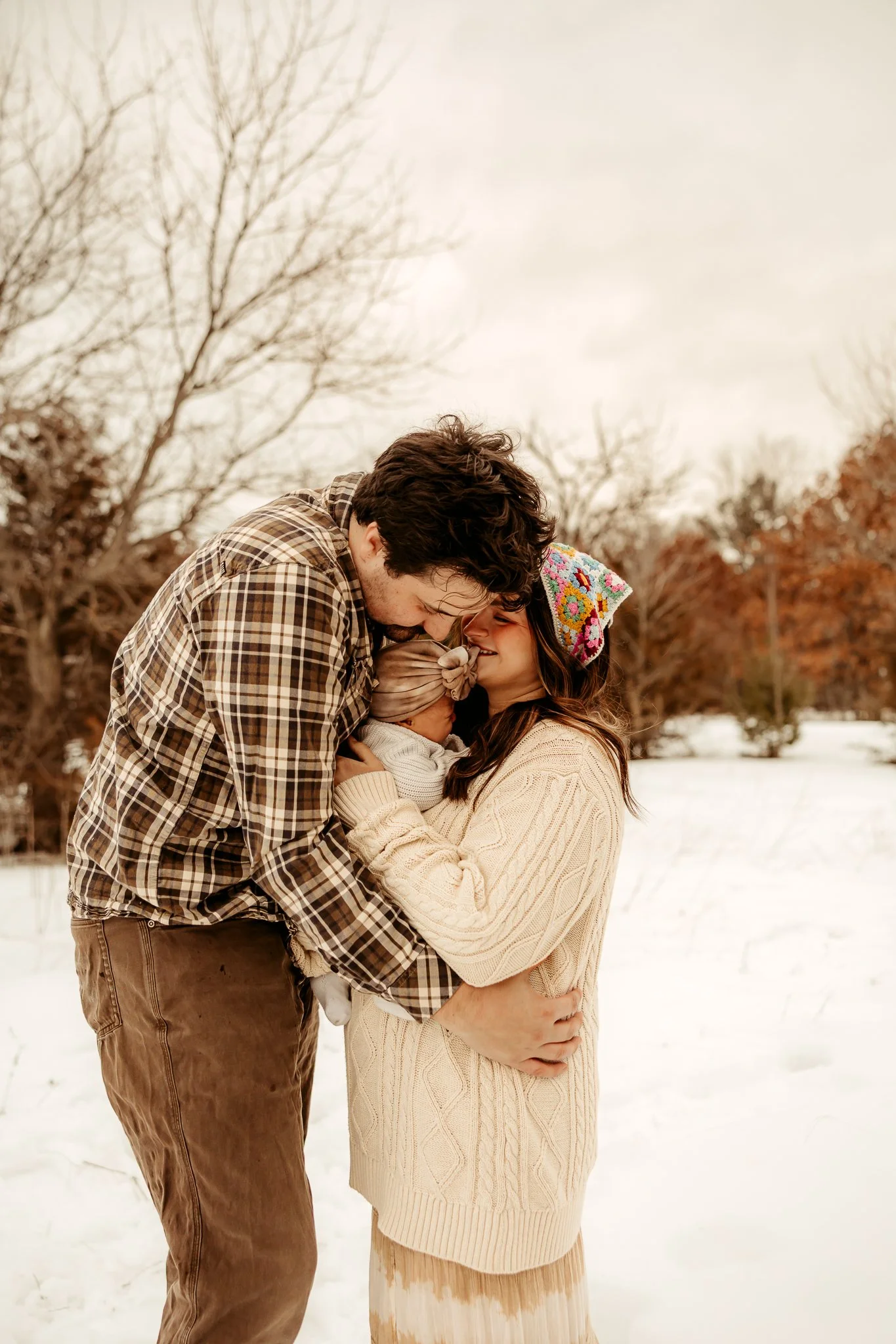A family of three in a winter landscape, with snow on the ground and leafless trees in the background. The man and woman are smiling and touching noses while holding their baby.