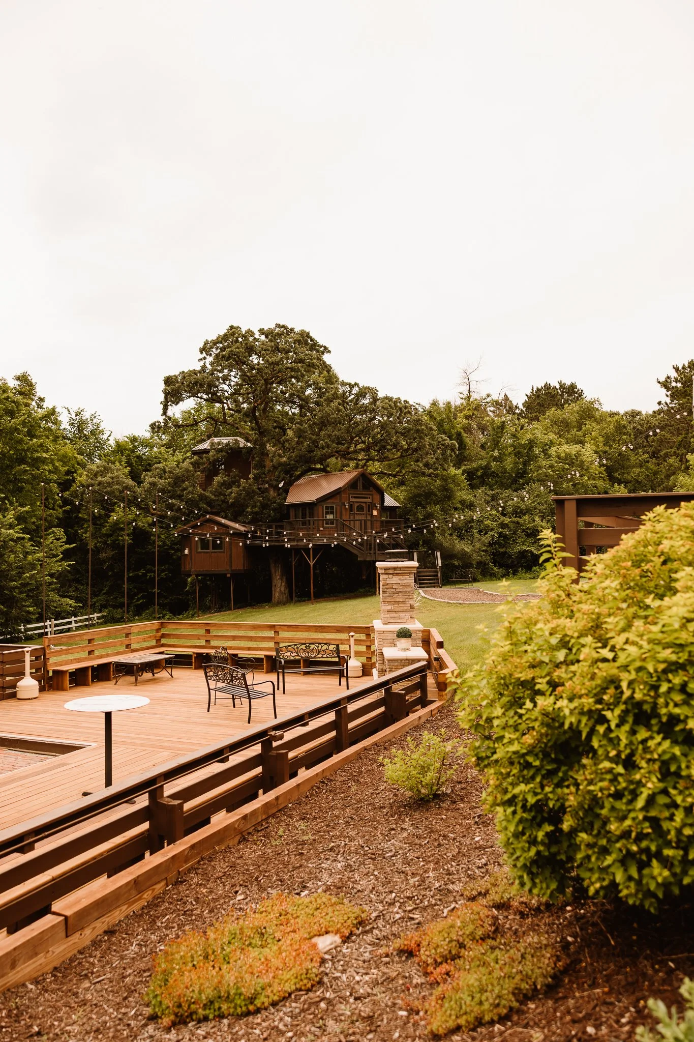 A backyard wooden deck with outdoor furniture, string lights, and a treehouse in the background surrounded by trees.