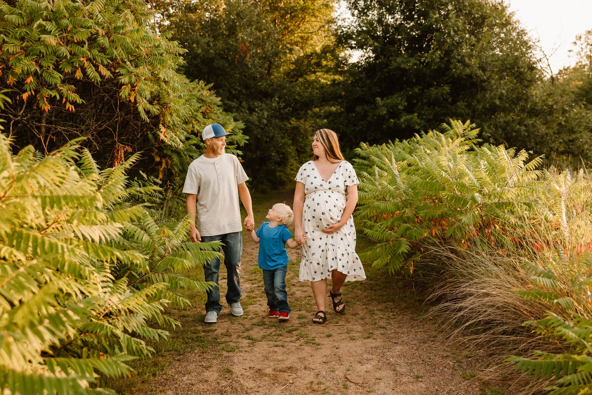 A family of four, including a pregnant woman, a man, a young boy, and a girl, holding hands and walking on a dirt path through a lush green forest during sunset.