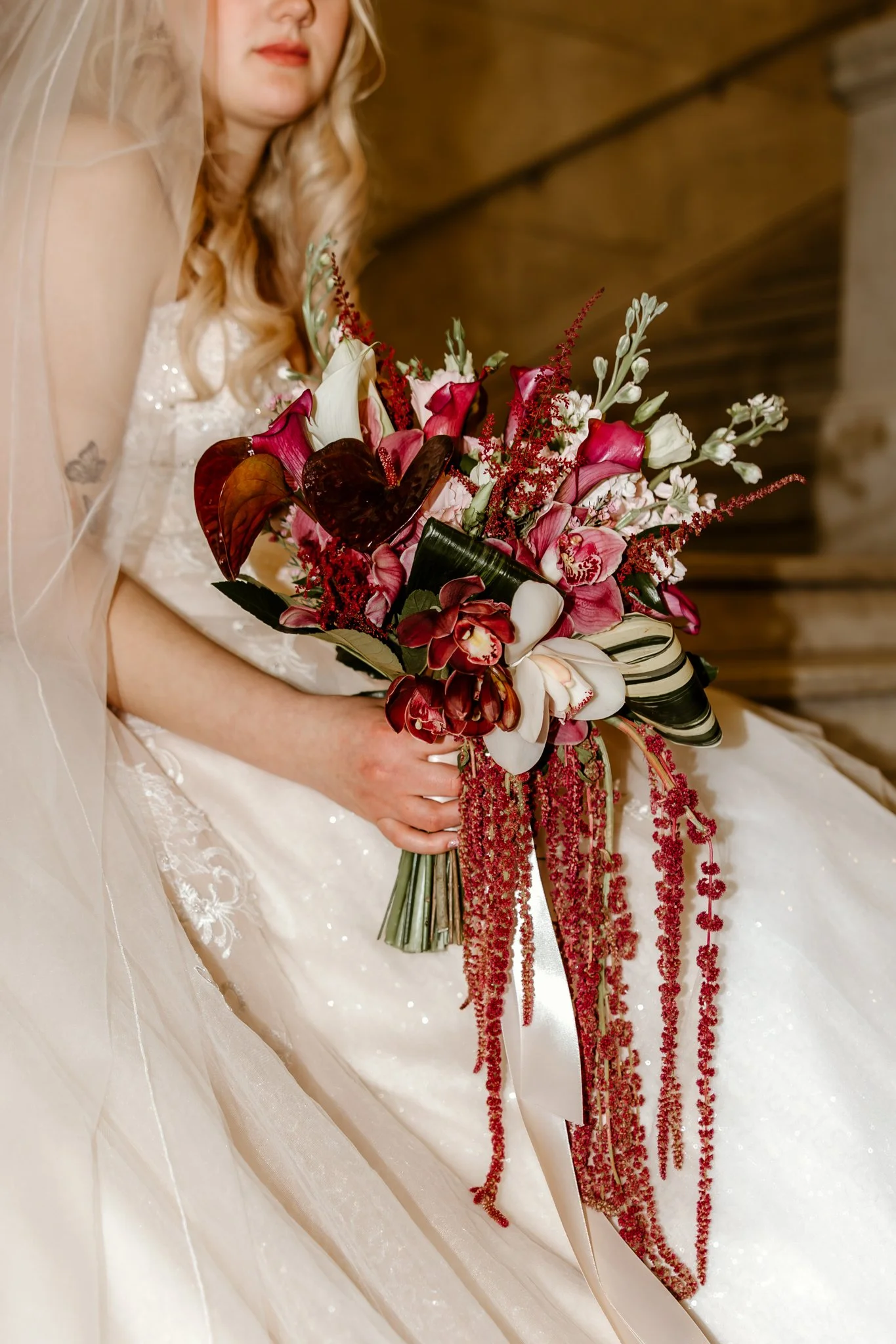 A bride holding a large bouquet of pink, white, and red flowers with greenery, sitting in a white wedding dress with lace details.