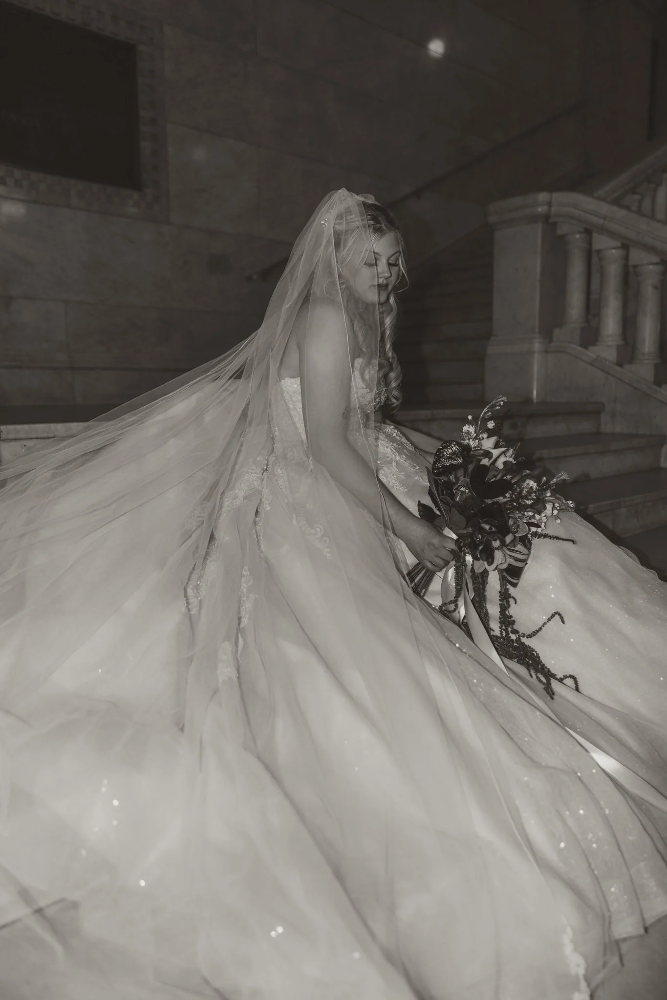 Black and white photo of a bride sitting on stairs holding a bouquet of flowers, wearing a wedding dress and veil.