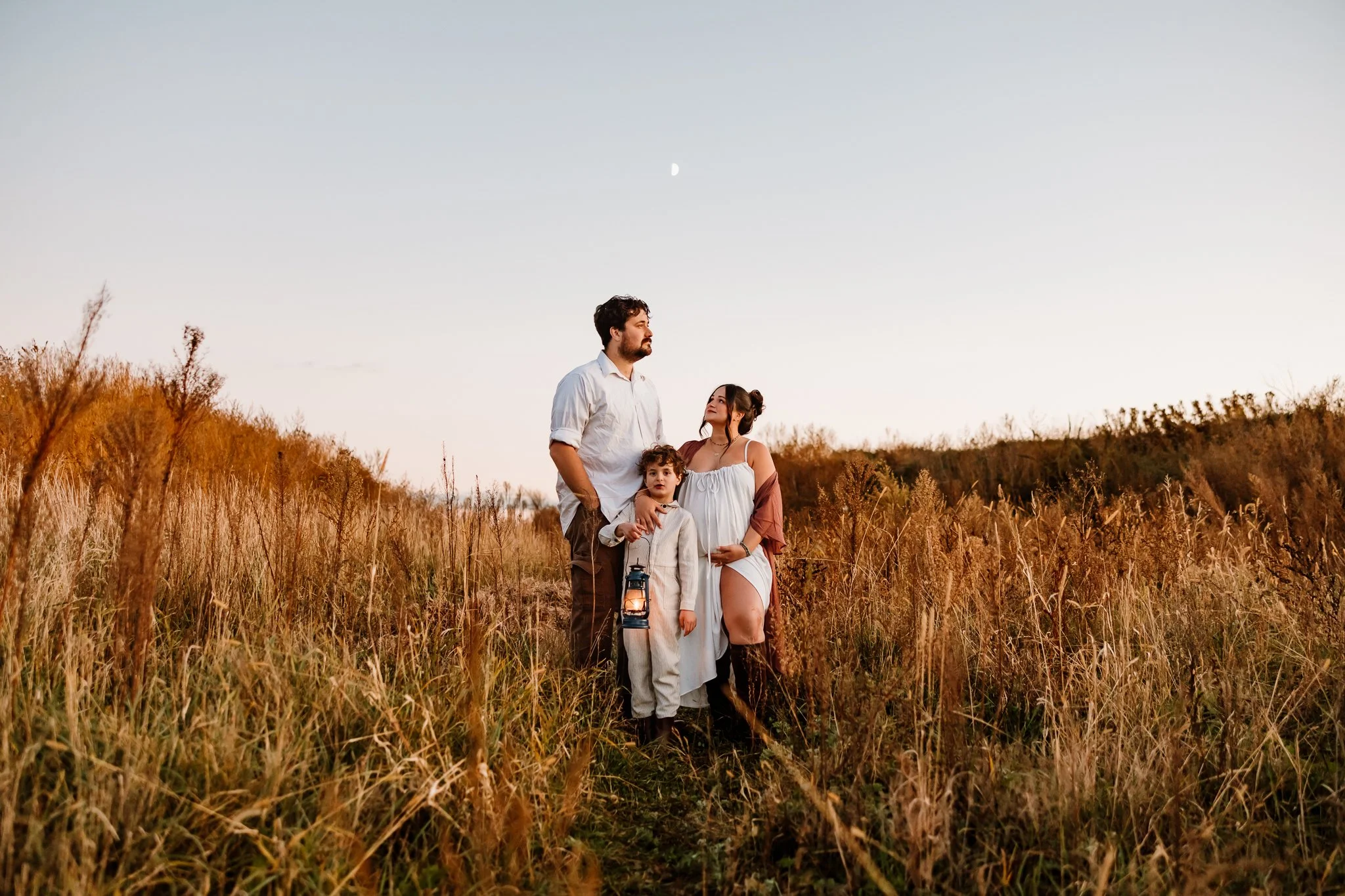 A family of three stands in a grassy field during sunset, with the moon visible in the sky. The father, mother, and young son look at each other and the surroundings, with the father holding a lantern.