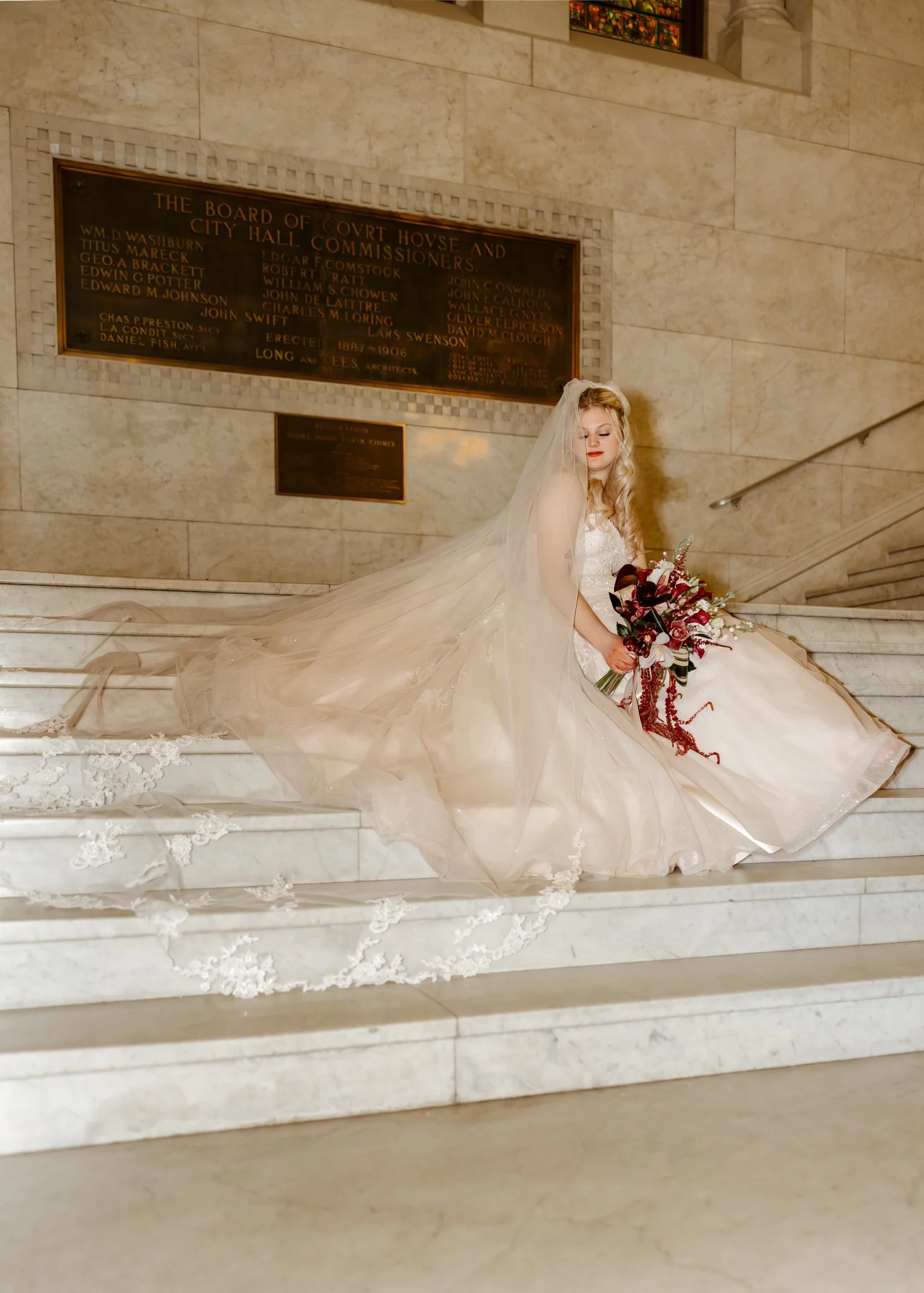 A bride in a white wedding gown with a long veil sitting on marble stairs inside a building, holding a bouquet of dark red and white flowers.