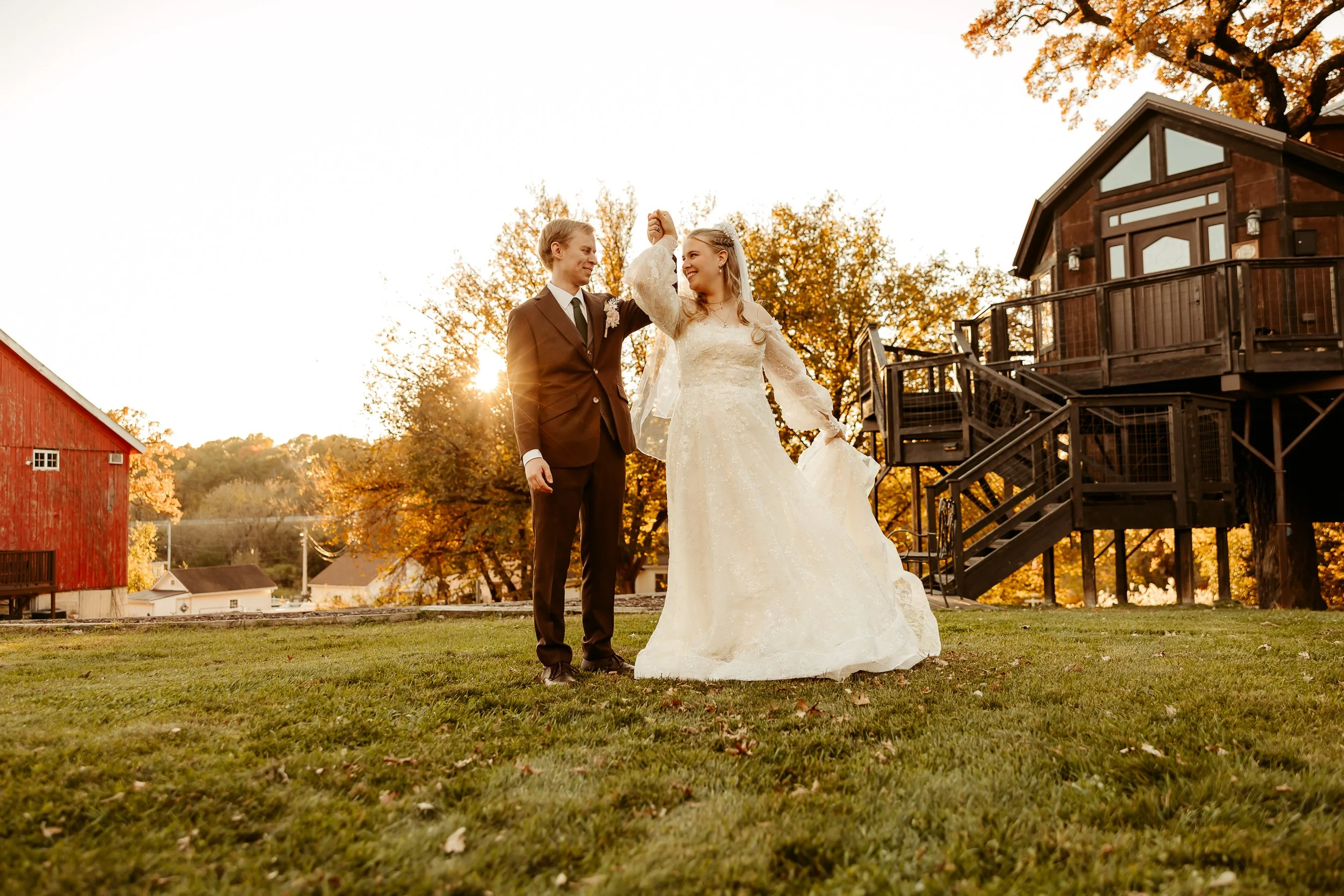 A bride and groom dancing outdoors during sunset, with the groom in a dark suit and the bride in a white wedding dress, on a green lawn with trees and buildings in the background. Minnesota wedding photographer at home glen farm 
