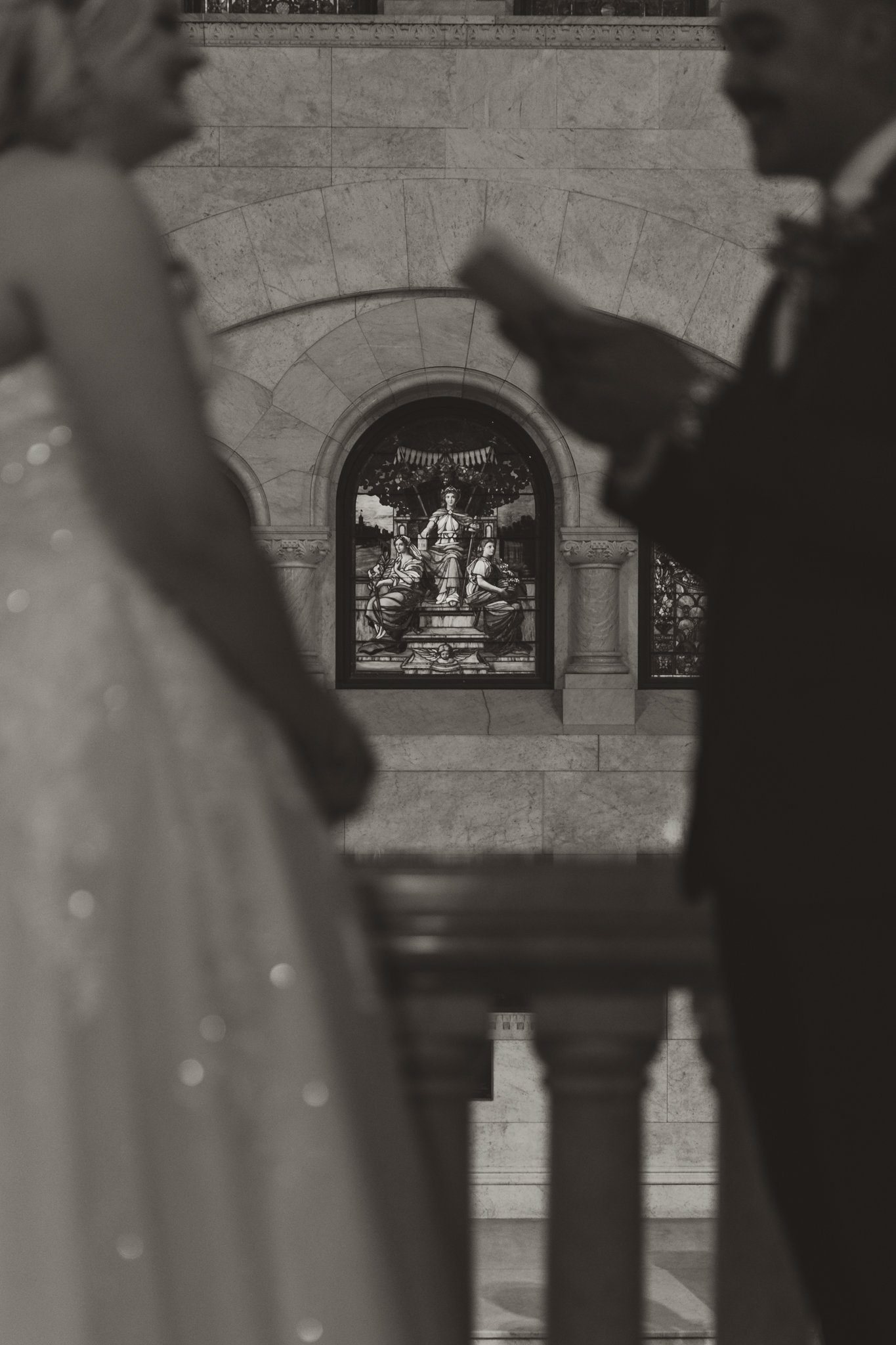 Statue of a woman in wedding dress with two men in suits, viewed through wrought iron railing, inside a church with stained glass window in the background.