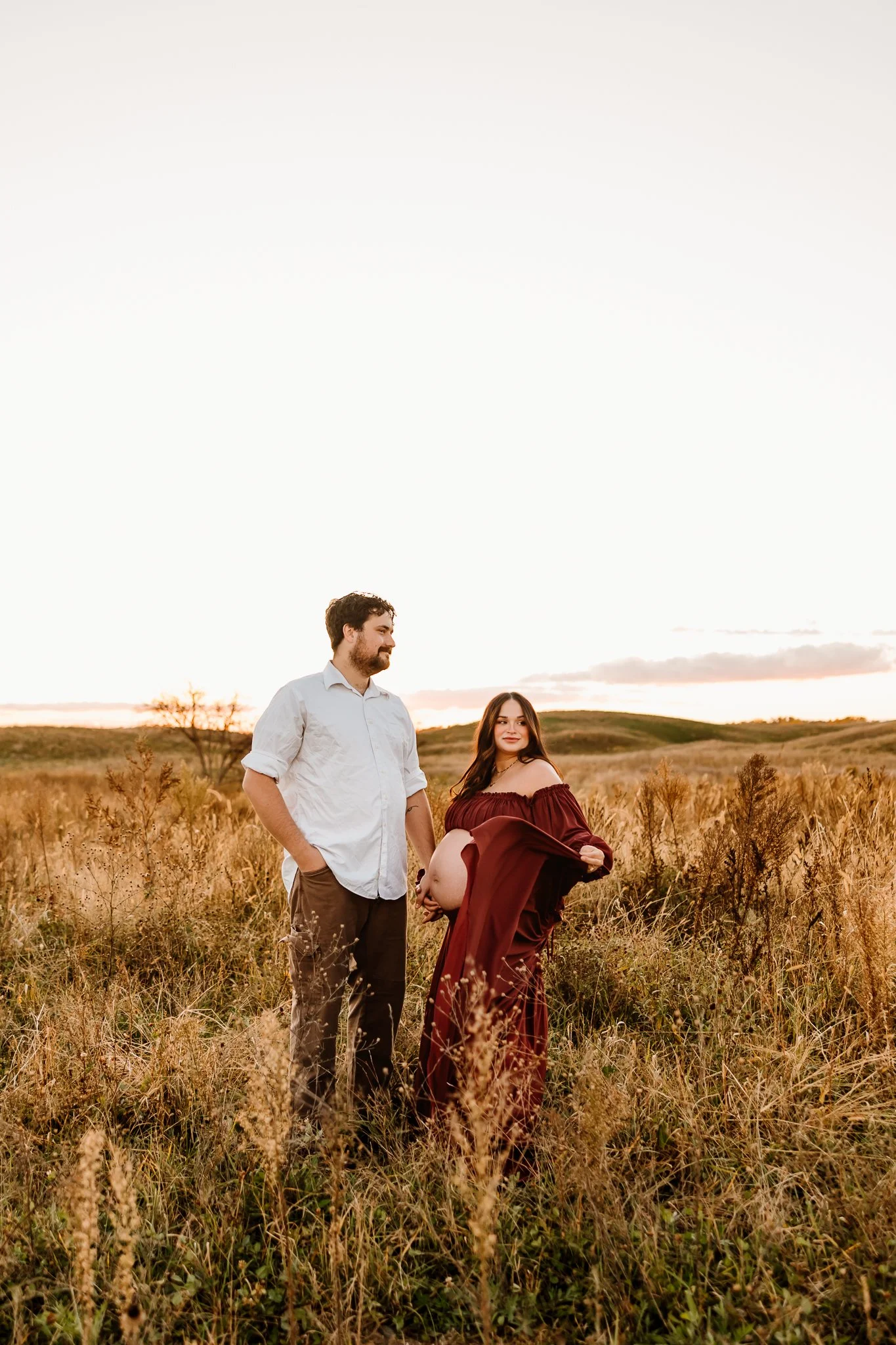 A pregnant woman in a flowing maroon dress standing in a field at sunset, holding her belly, with a man in a white shirt and brown pants standing beside her, both looking at each other.