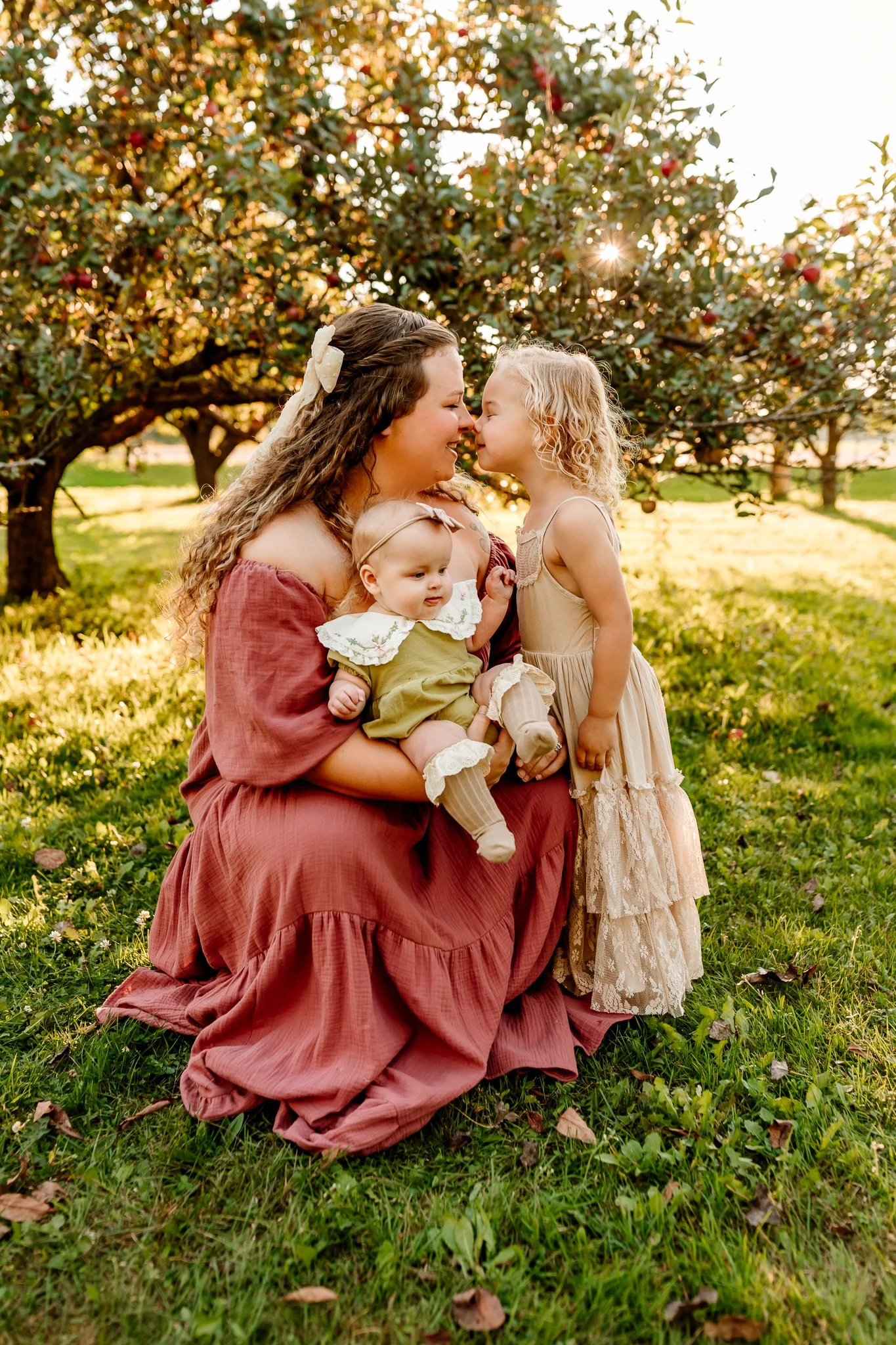 A woman dressed in a mauve off-shoulder dress sitting on the grass in an orchard with two young girls, touching noses, and smiling, during sunset.