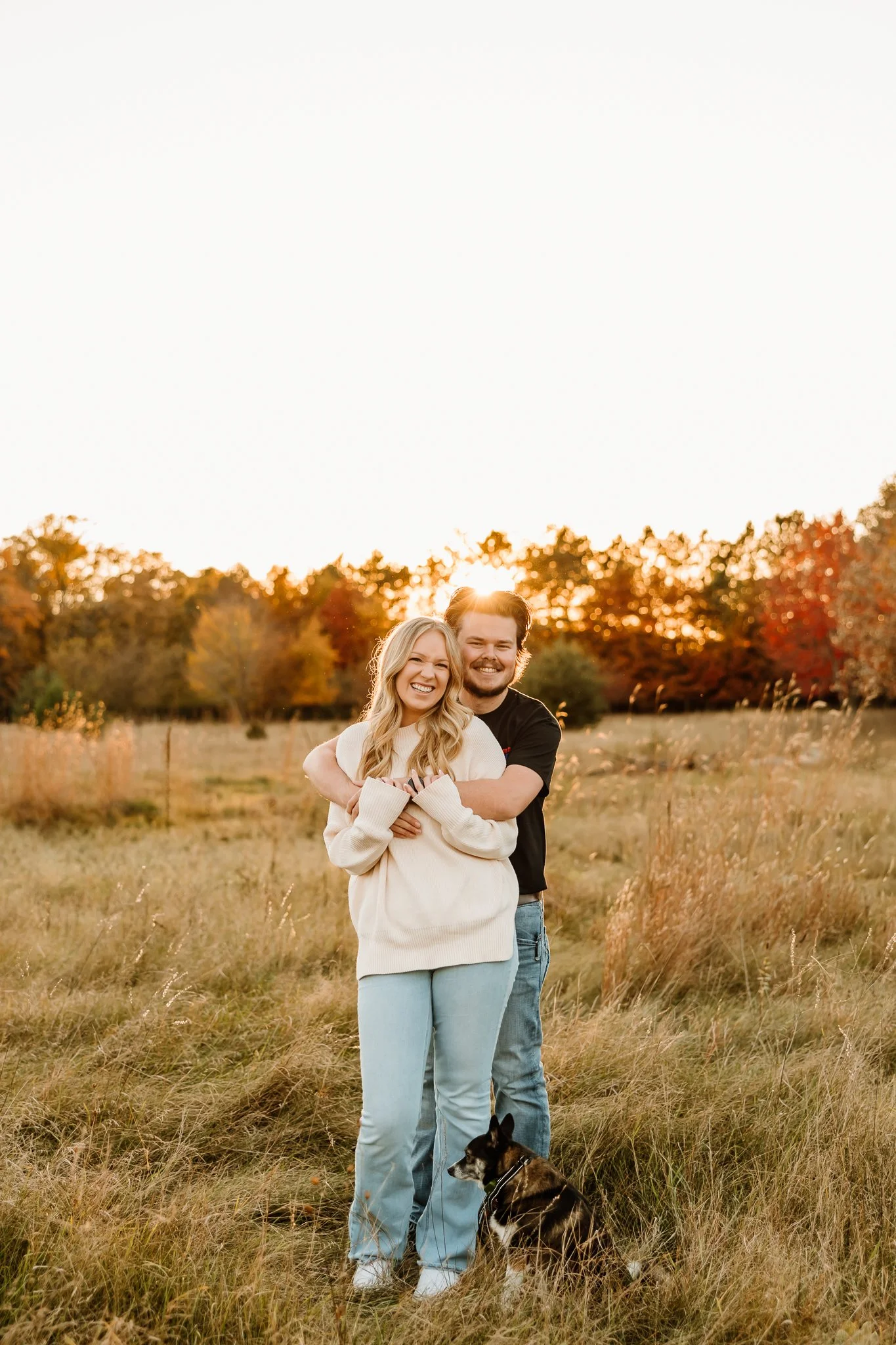 A couple hugging and smiling in a grassy field during autumn, with a small black and tan dog sitting in front of them and trees with fall foliage in the background at sunset.
