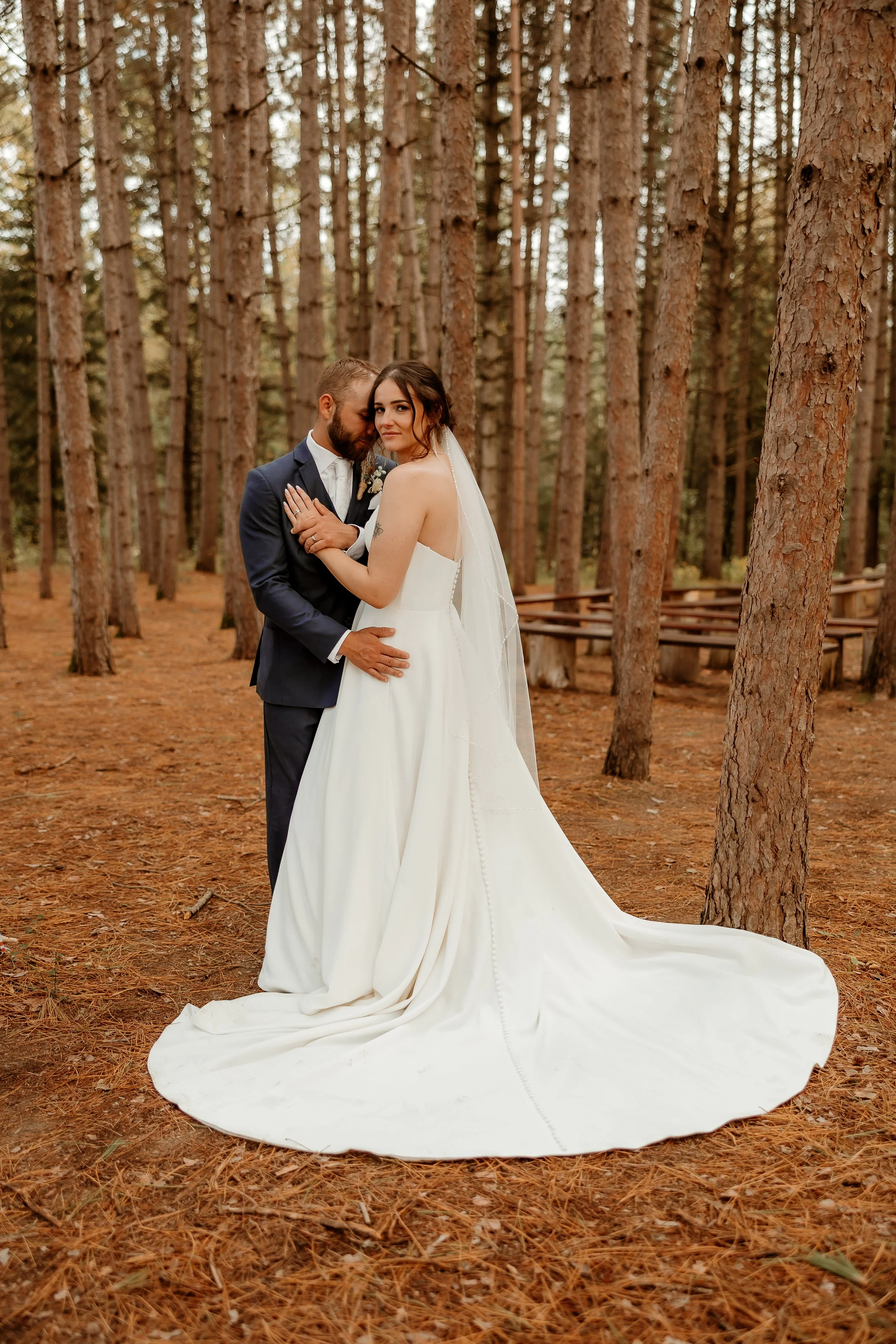 A bride and groom embrace in a forest with tall pine trees, the bride in a white wedding gown and veil, and the groom in a dark suit, during a wedding photoshoot.
