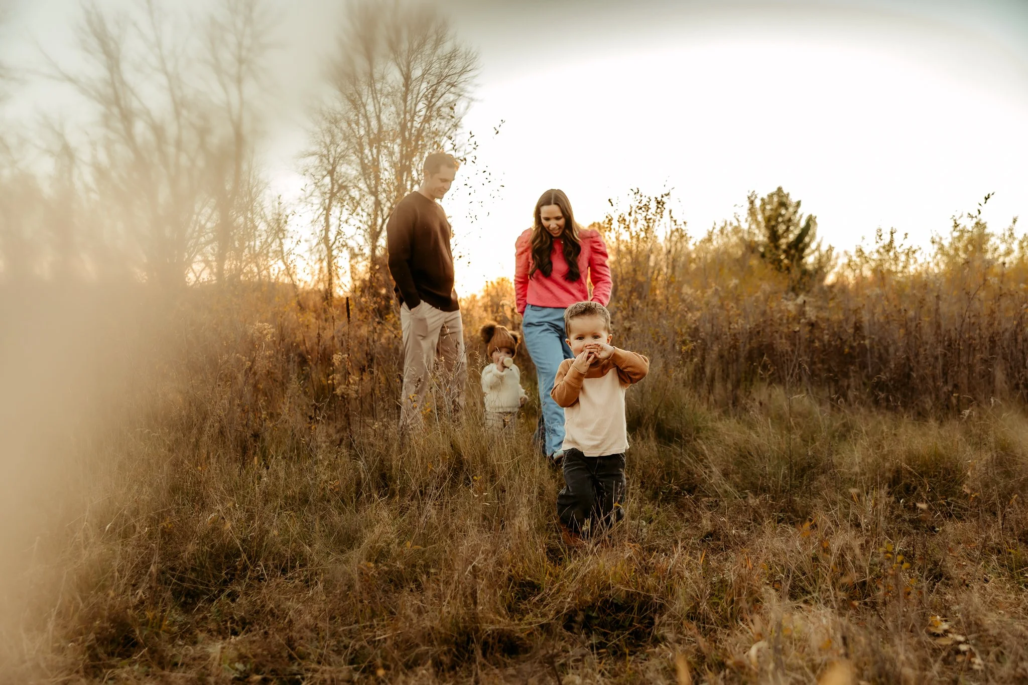 A family of four walking outdoors in a grassy field during sunset. The young boy at front is smiling and holding his hands near his mouth. The girl behind him is wearing a white sweater and has her hair in buns. The woman has long dark hair and a pin