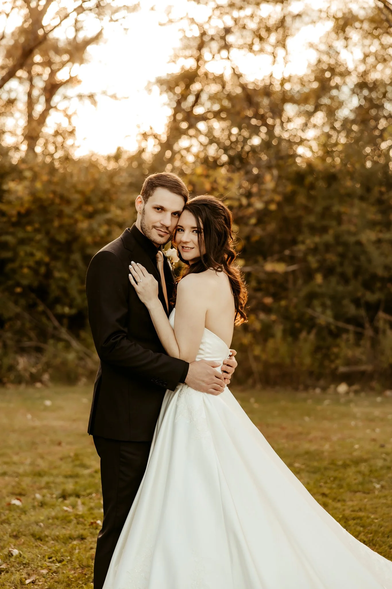 A newlywed couple stands closely together outdoors during sunset, with trees and golden light in the background. The bride is in a white strapless wedding gown, and the groom is wearing a black suit with a white shirt and tie. They are smiling and em